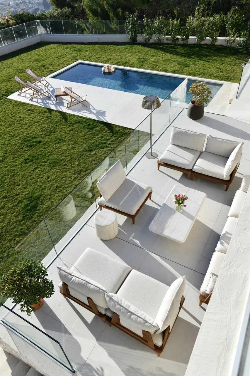 Terrasse avec canapé, fauteuils, table basse avec des fleurs, lampe sur pied, piscine rectangulaire et pelouse verte entourée de murs et de végétation.