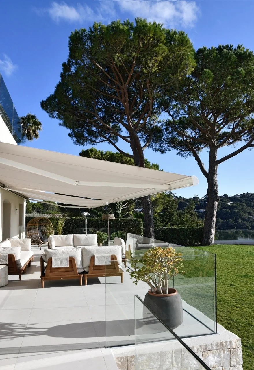 Terrasse extérieure élégante avec mobilier blanc, grande ombrière, pot de plante, et grands pins sous un ciel bleu.