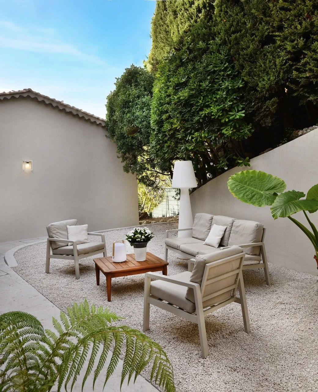 Espace extérieur avec mobilier de salon en bois et coussins beige, entouré de végétation luxuriante et une allée en gravier. Ciel clair avec quelques nuages.