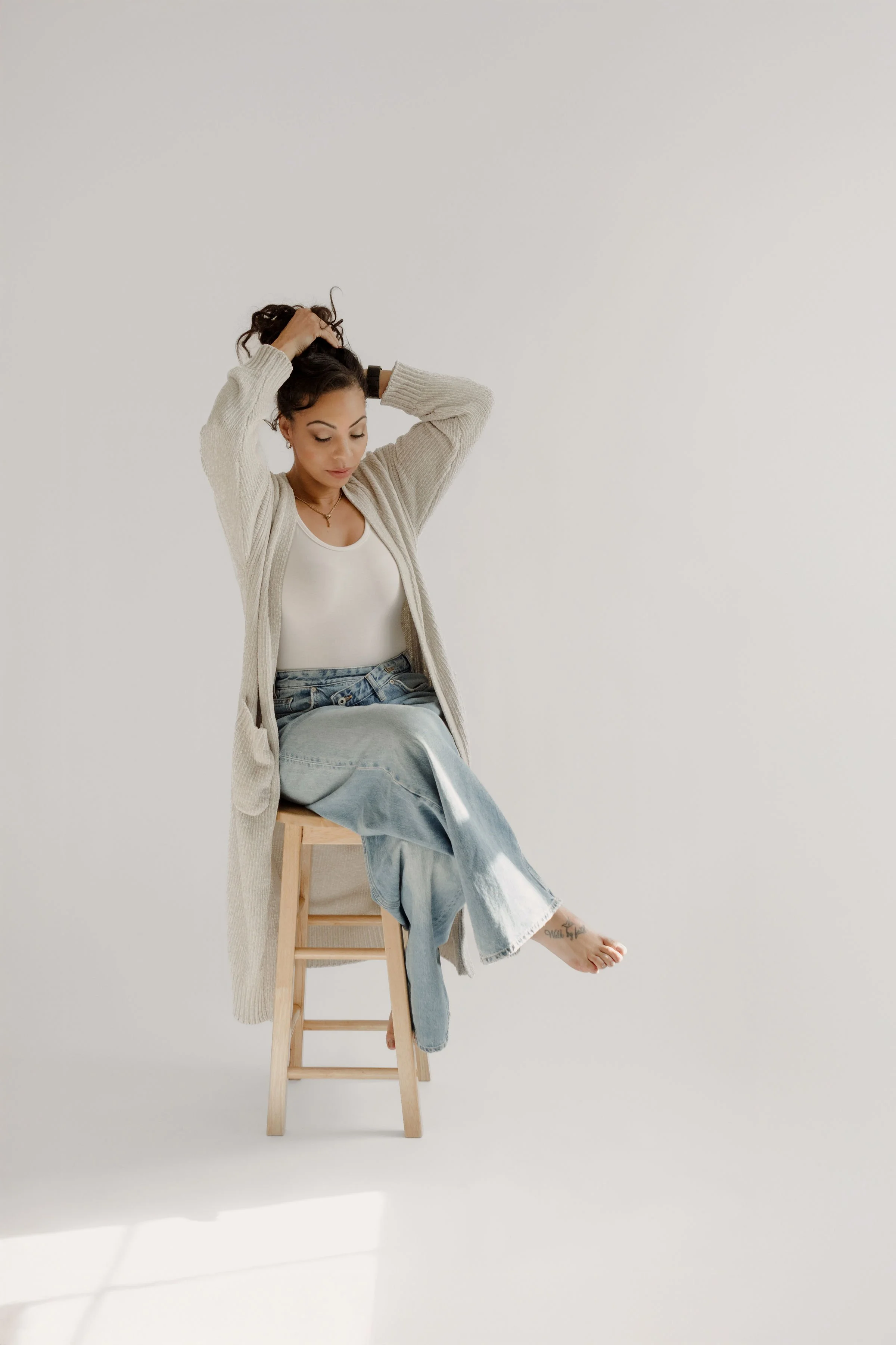 A woman with dark curly hair styled in a bun, sitting on a wooden stool against a plain white background. She is wearing a beige cardigan, white top, and light blue jeans. She is lifting her arms to adjust her hair, with one leg crossed over the other, and her foot shows a small tattoo.