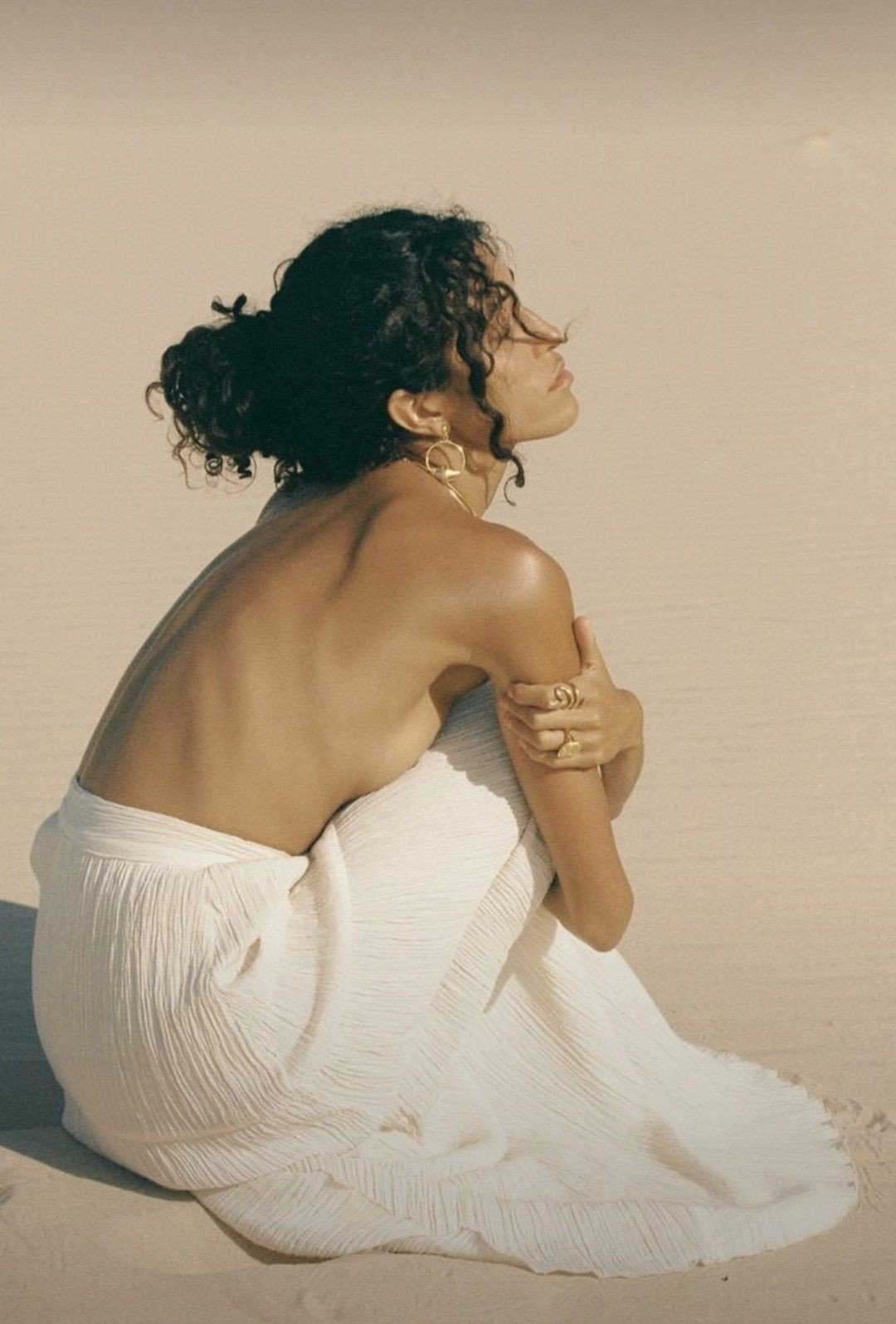 A woman with dark, curly hair in a bun, wearing gold jewelry, sitting on the floor with her knees drawn up, wrapped in a white pleated fabric, against a plain beige background.