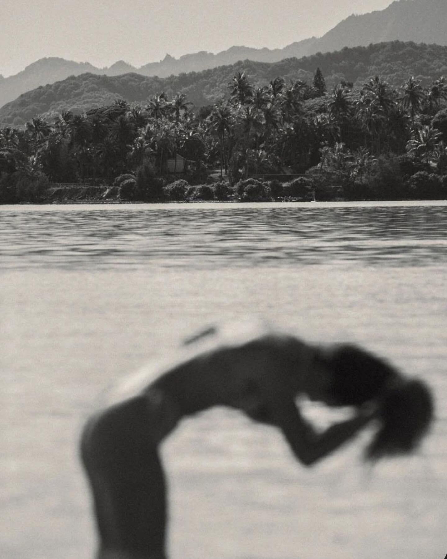 Black and white photo of a person doing a backbend on a sandy beach with a forested hill and mountains in the background.