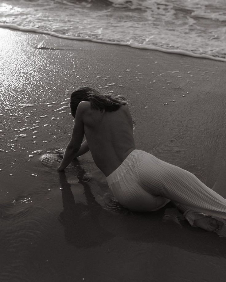 A woman in white pants kneeling on wet sand at the edge of the ocean, facing away from the camera with her long hair flowing.
