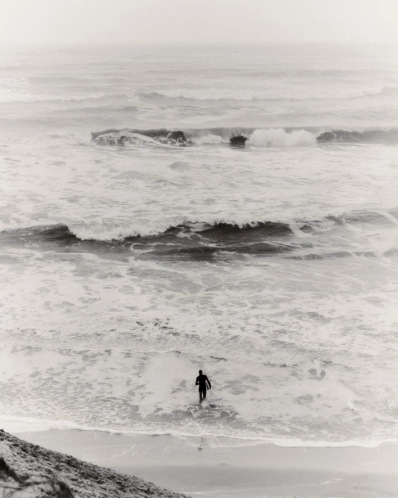 A person standing in the ocean waves, with large waves and overcast sky in the background, black and white photo.
