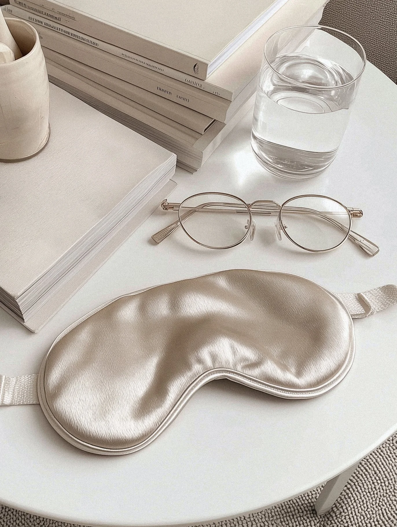 A white table with a silver sleep mask, round eyeglasses, a glass of water, some stacked books, a closed notebook, and a ceramic cup.