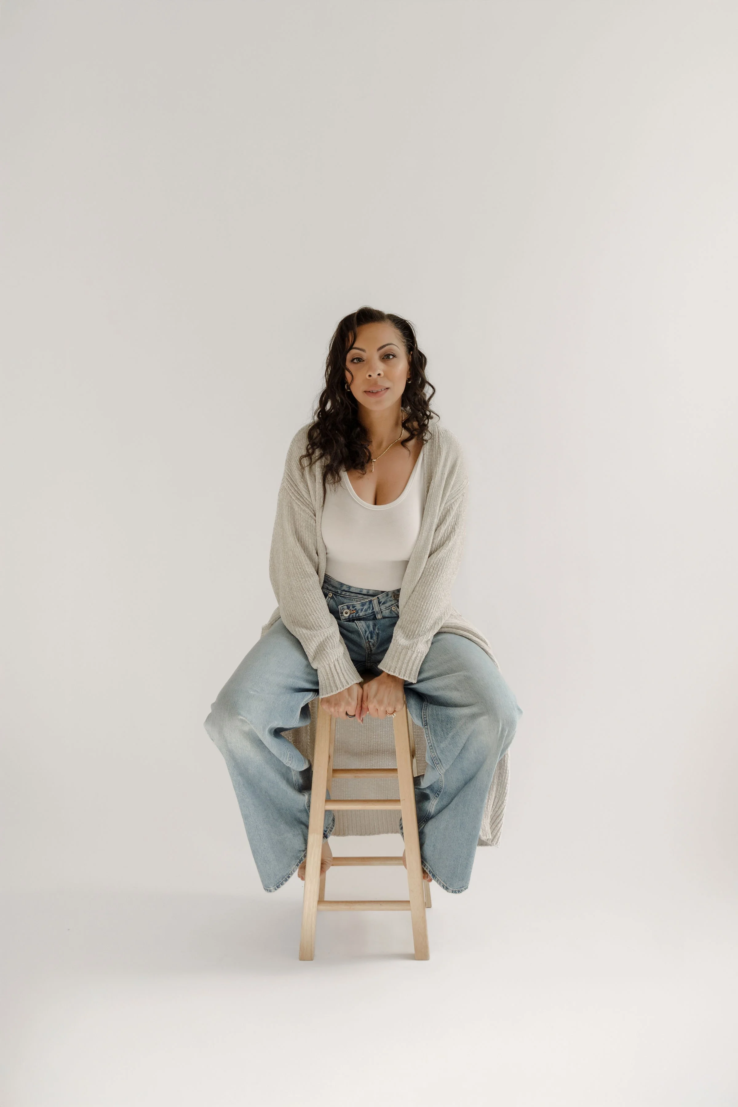 A woman with curly dark hair sitting on a wooden stool against a plain white background, wearing a white top, light blue jeans, and a beige cardigan.
