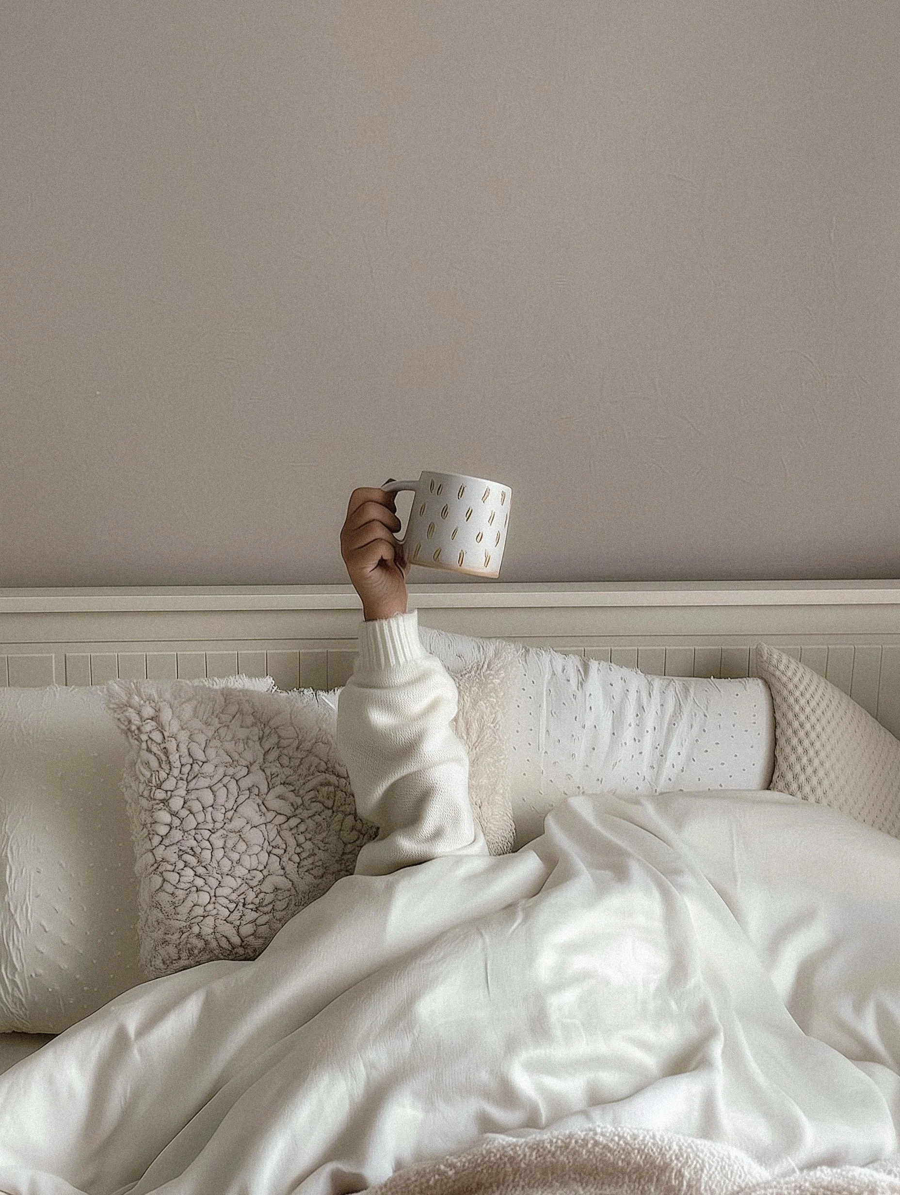 Person lying in bed, raising an hand with a mug that has a pattern of gold streaks, in a cozy, neutral-colored bedroom.
