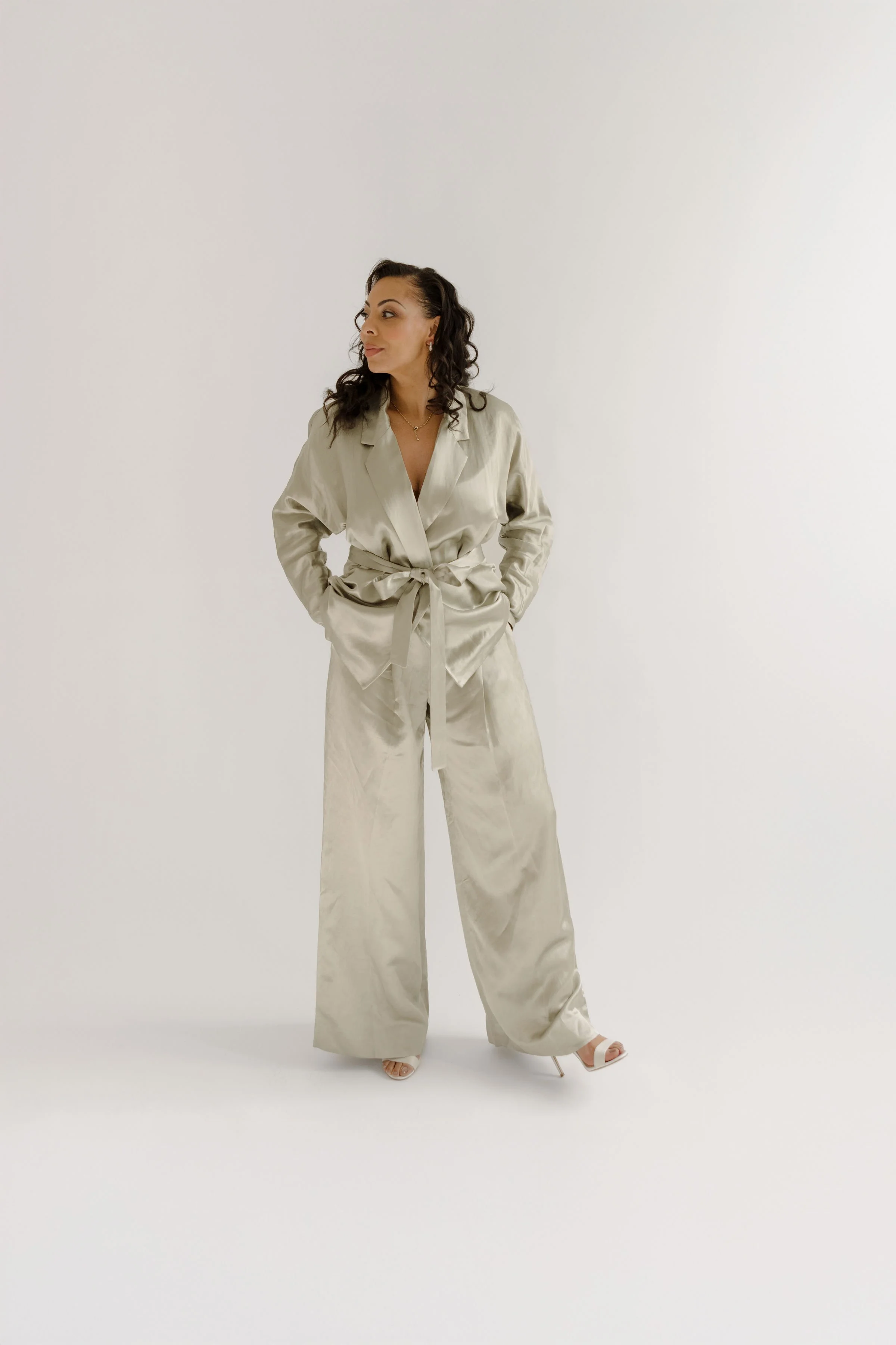A woman in a cream-colored silk suit with wide-leg pants and a tie-front blazer, standing against a plain white background, looking to her left.