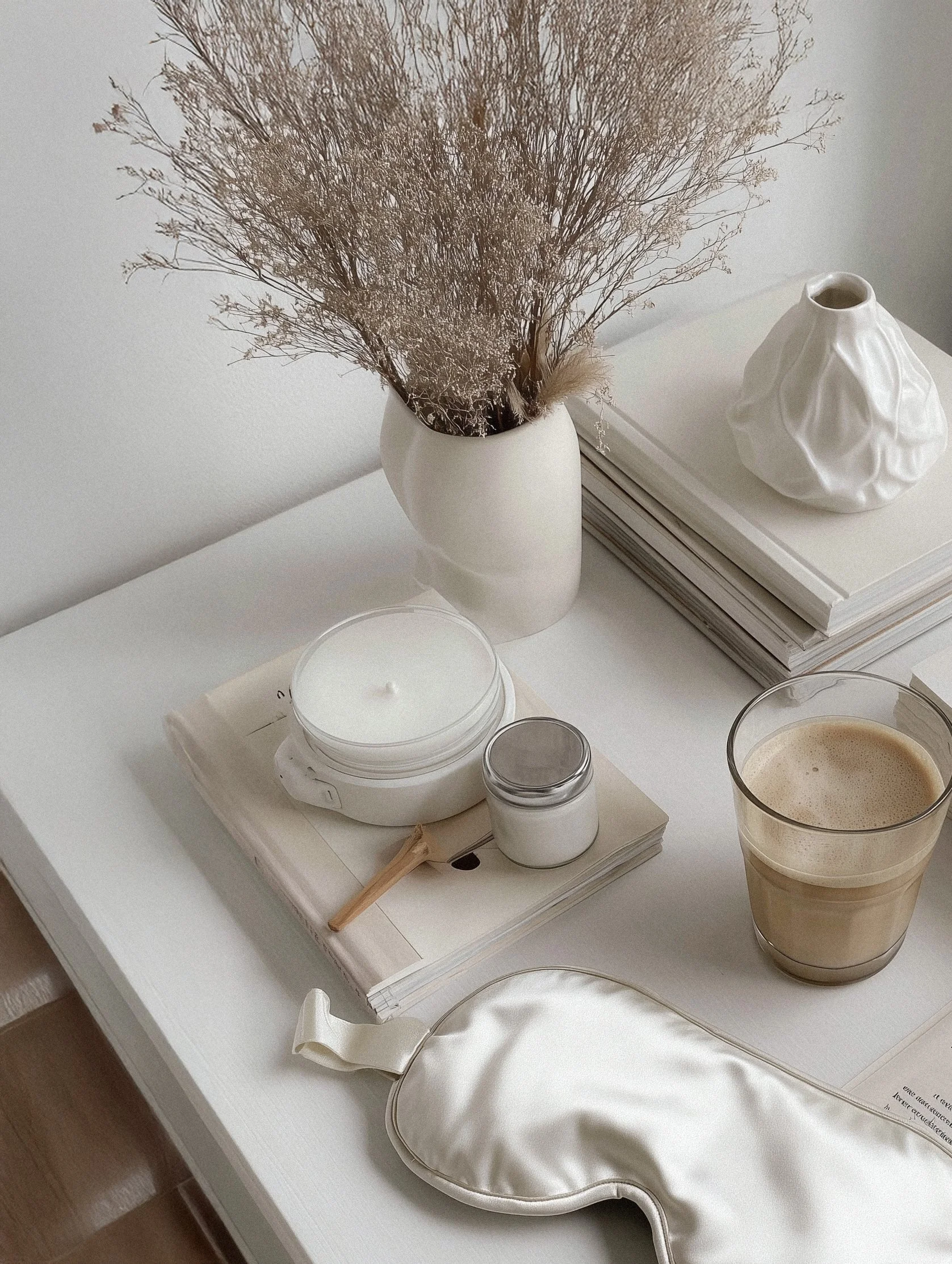 A white table with a vase of dried beige flowers, a white ceramic decorative object, a stack of books, a candle on a book, a small jar, a glass of coffee, a silky eye mask, and some magazines.