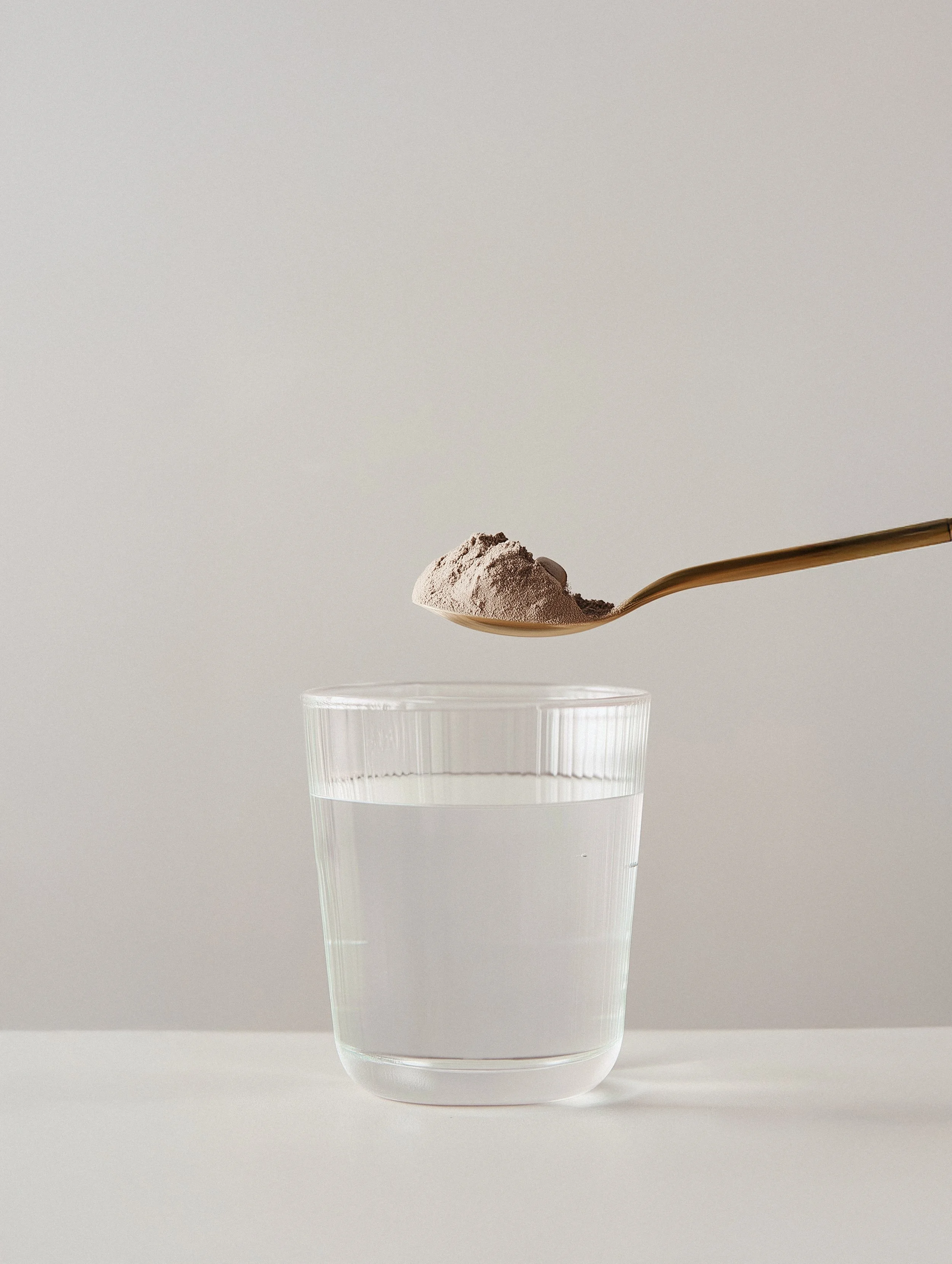 A transparent glass of water with a spoonful of gray powder above it.