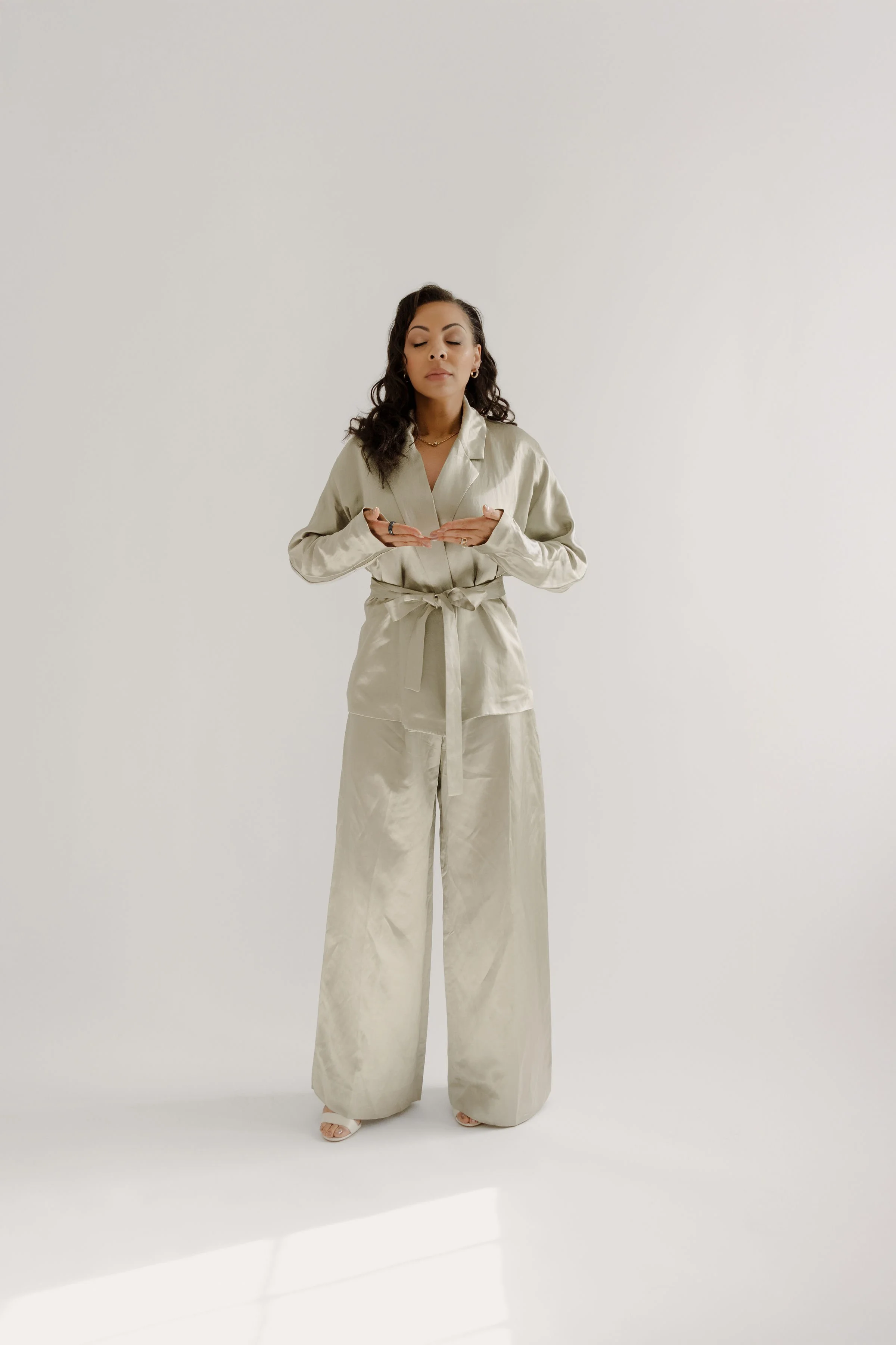 Woman in cream-colored satin pajamas with wide-leg pants and a tied waist, standing against a plain white background, with eyes closed and hands in a prayer position.