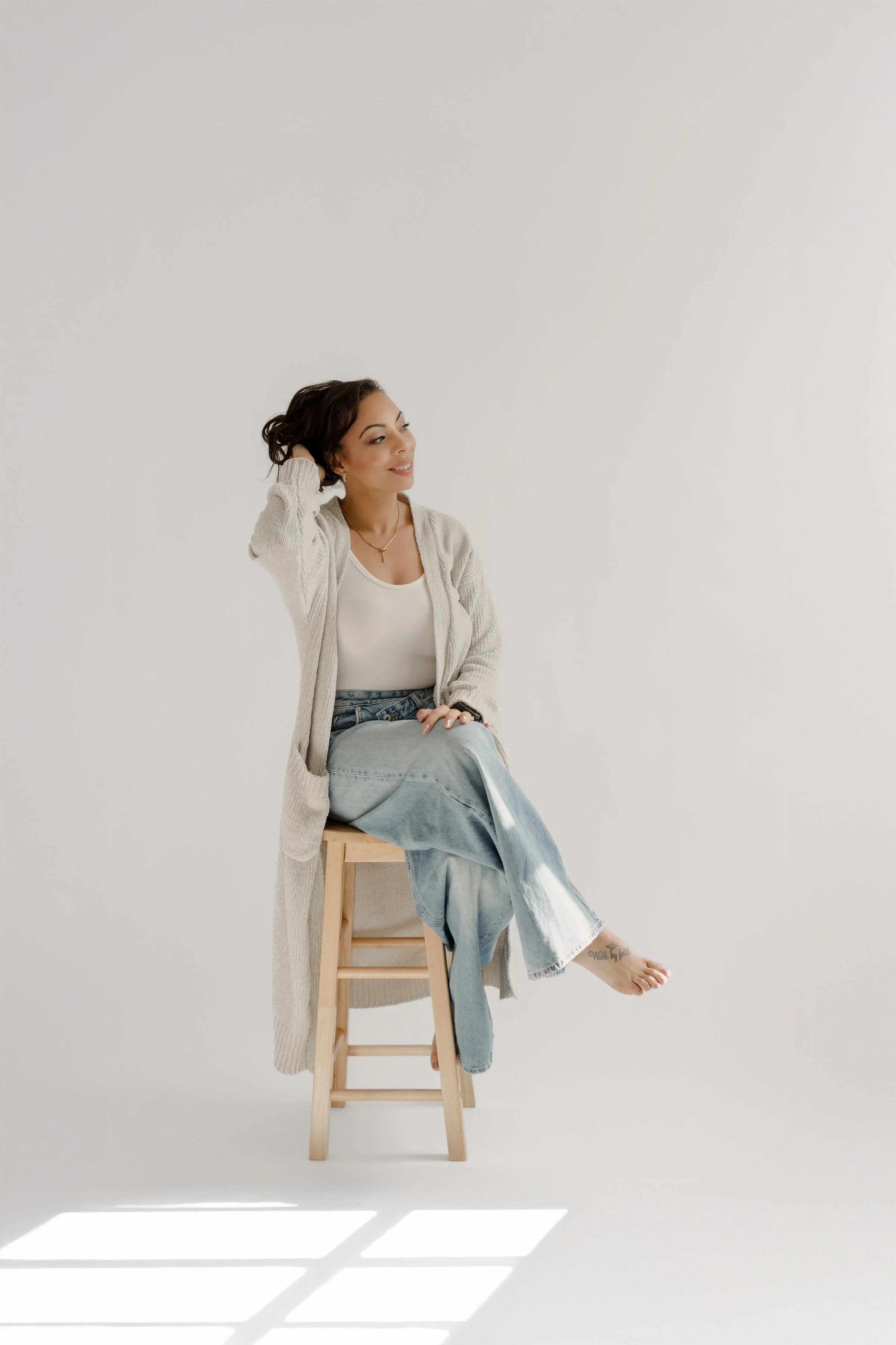 A woman sitting on a wooden stool with one leg crossed over the other, smiling and looking to the side, wearing a beige cardigan, white shirt, and light blue jeans, against a plain white wall with sunlight casting shadows.