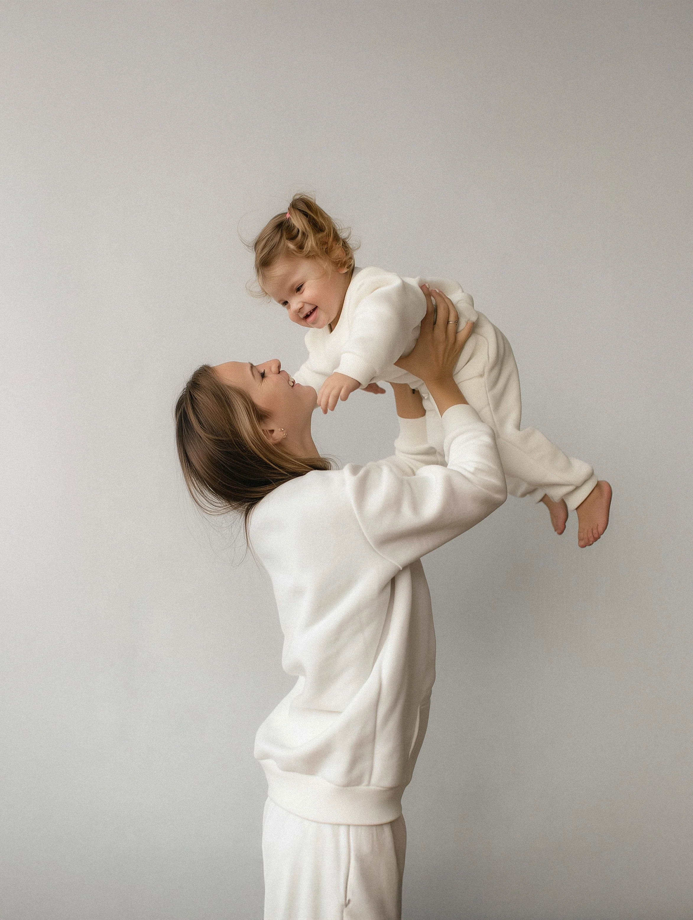 A woman lifting a young girl in the air, both smiling and looking at each other against a plain white background.