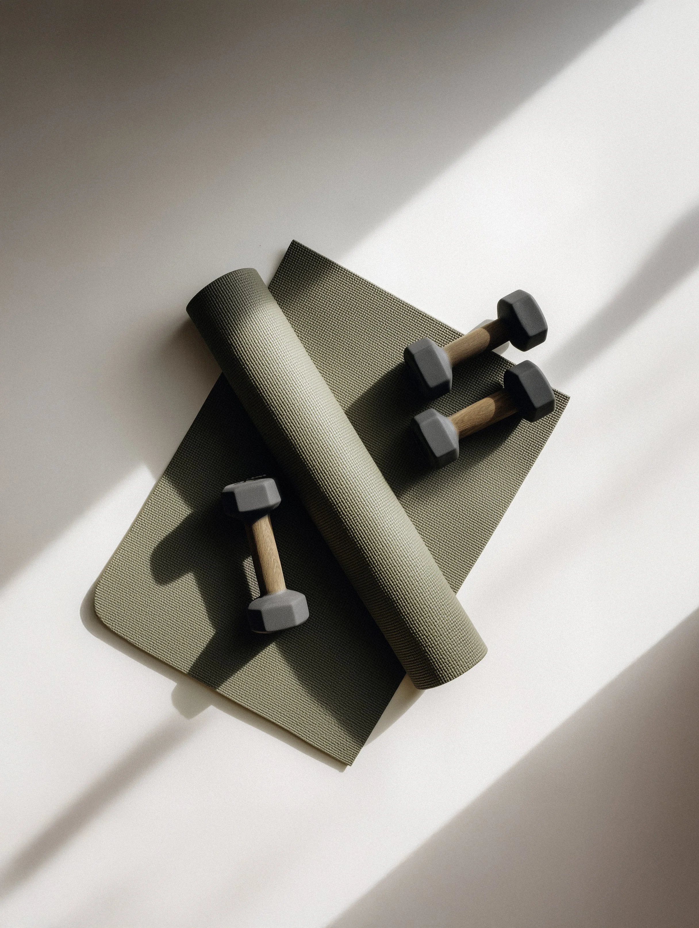 Green yoga mat, foam roller, and two dumbbells on a white surface with sunlight casting shadows.