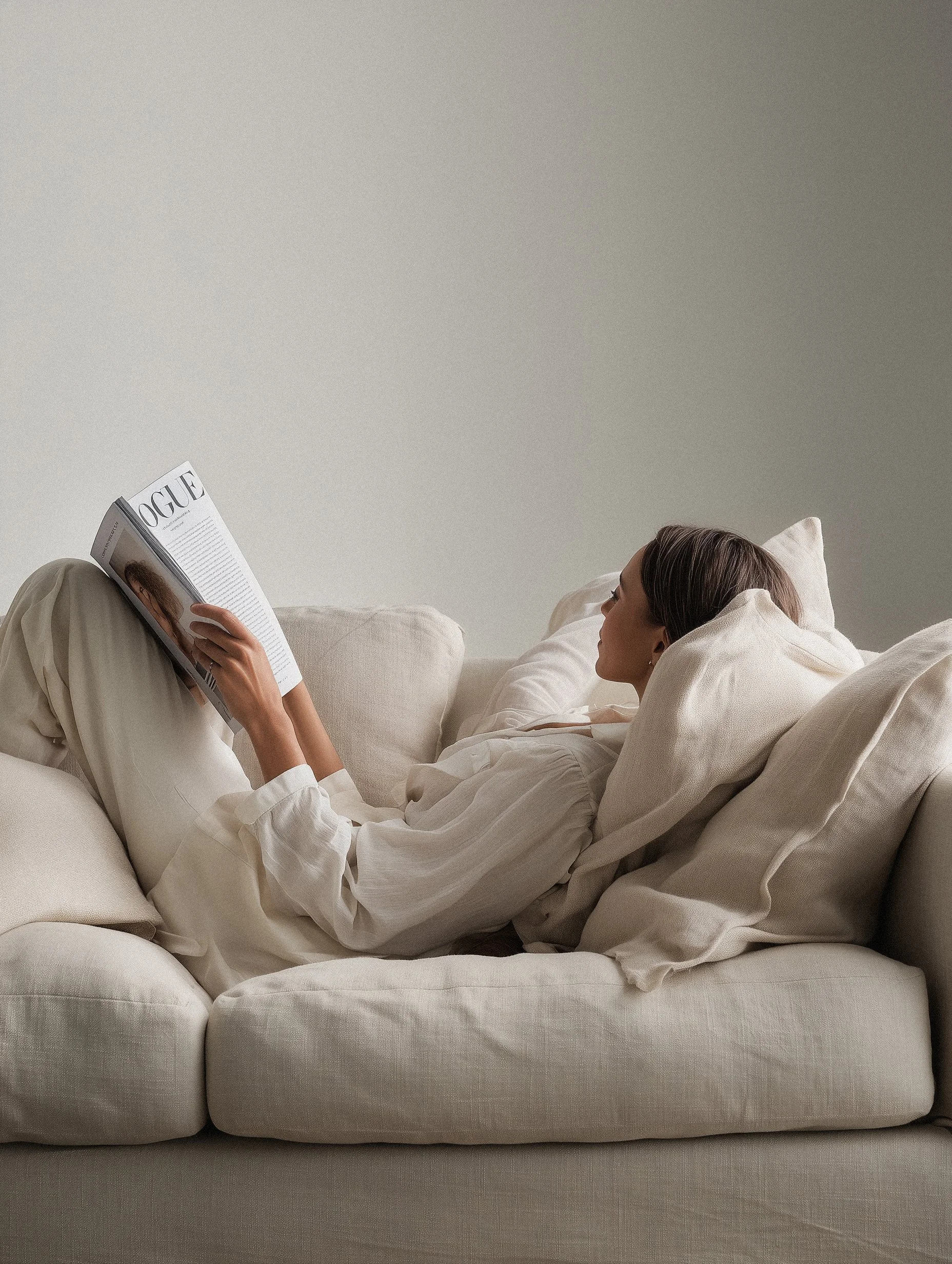 A woman lying on a white couch, reading a magazine titled 'VOGUE' in a minimalistic room with a plain wall.