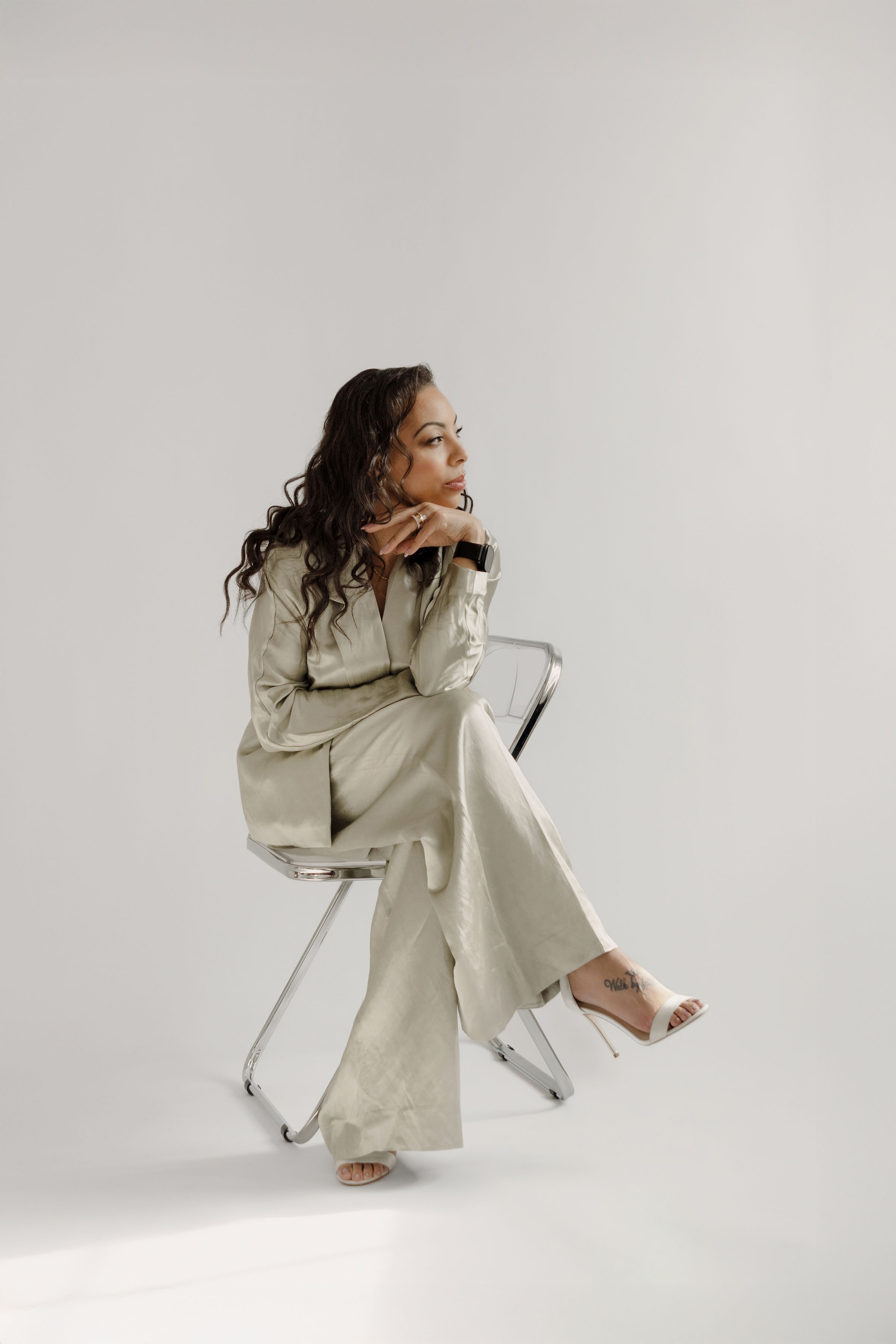 A woman with long curly dark hair sitting on a clear plastic chair with metal legs, wearing a cream-colored satin suit and white high-heeled sandals, looking to her right with her chin resting on her hand against a plain white background.