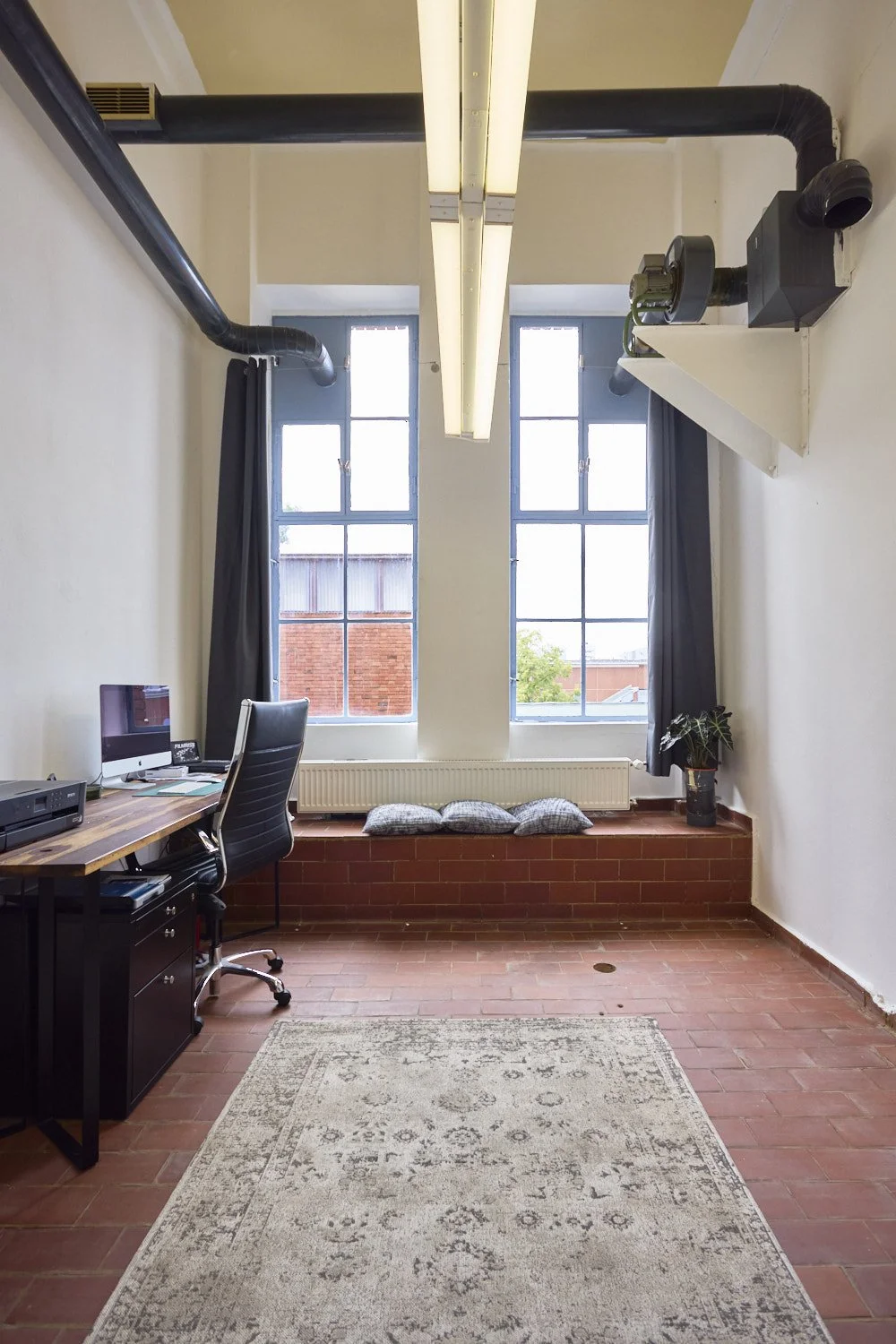 An office with two tall windows, a desk with an iMac and printer, a black office chair, a rug, pillows on a brick window seat, a potted plant, and overhead industrial pipes and lighting.