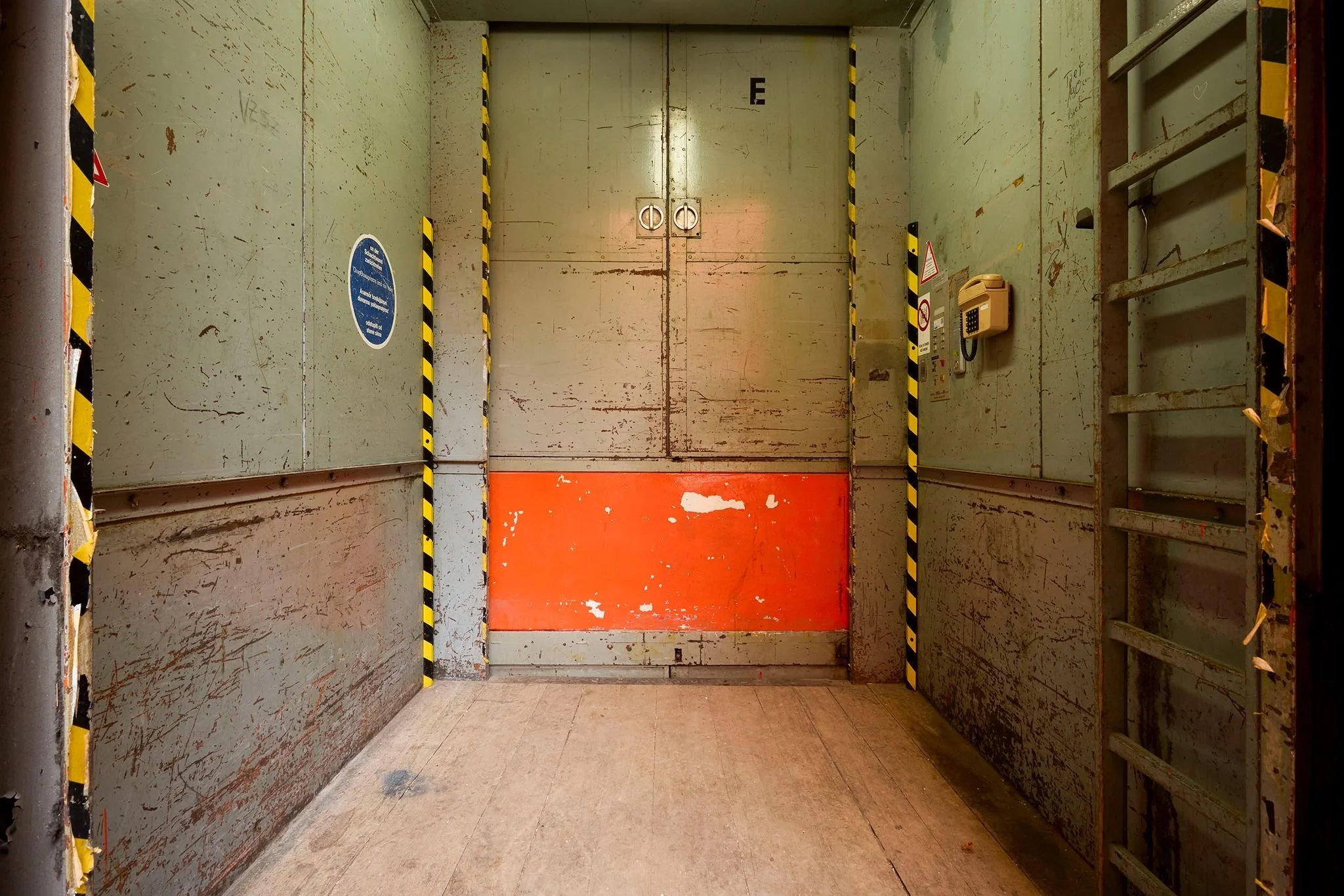 An elevator with closed metal doors, labeled with an E at the top and surrounded by black and yellow caution tape, with a red floor marking in front.