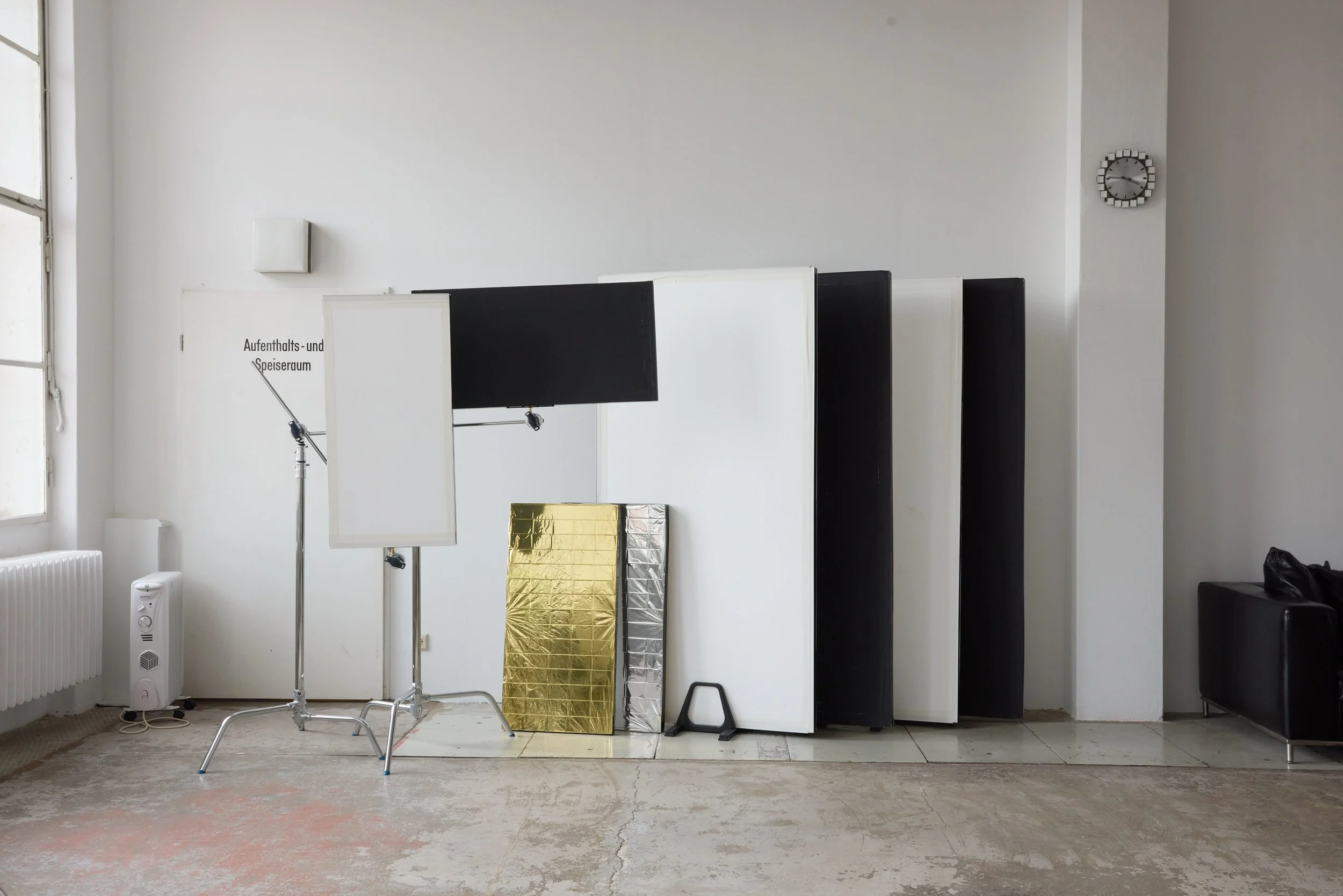 An empty room with a white wall, a window on the left, and a wall clock on the right. There are studio lighting setups, reflection panels, and gold reflective material on the floor.