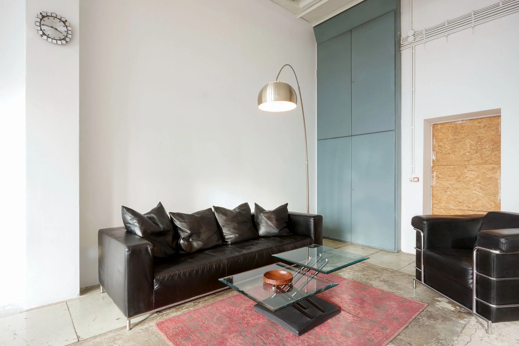 Modern living room with black leather sofa set, glass coffee table, and a red rug. There is a floor lamp with a metallic shade, a white wall with a clock, and a green sliding door with a panel. Part of a wooden panel is visible on the right side.