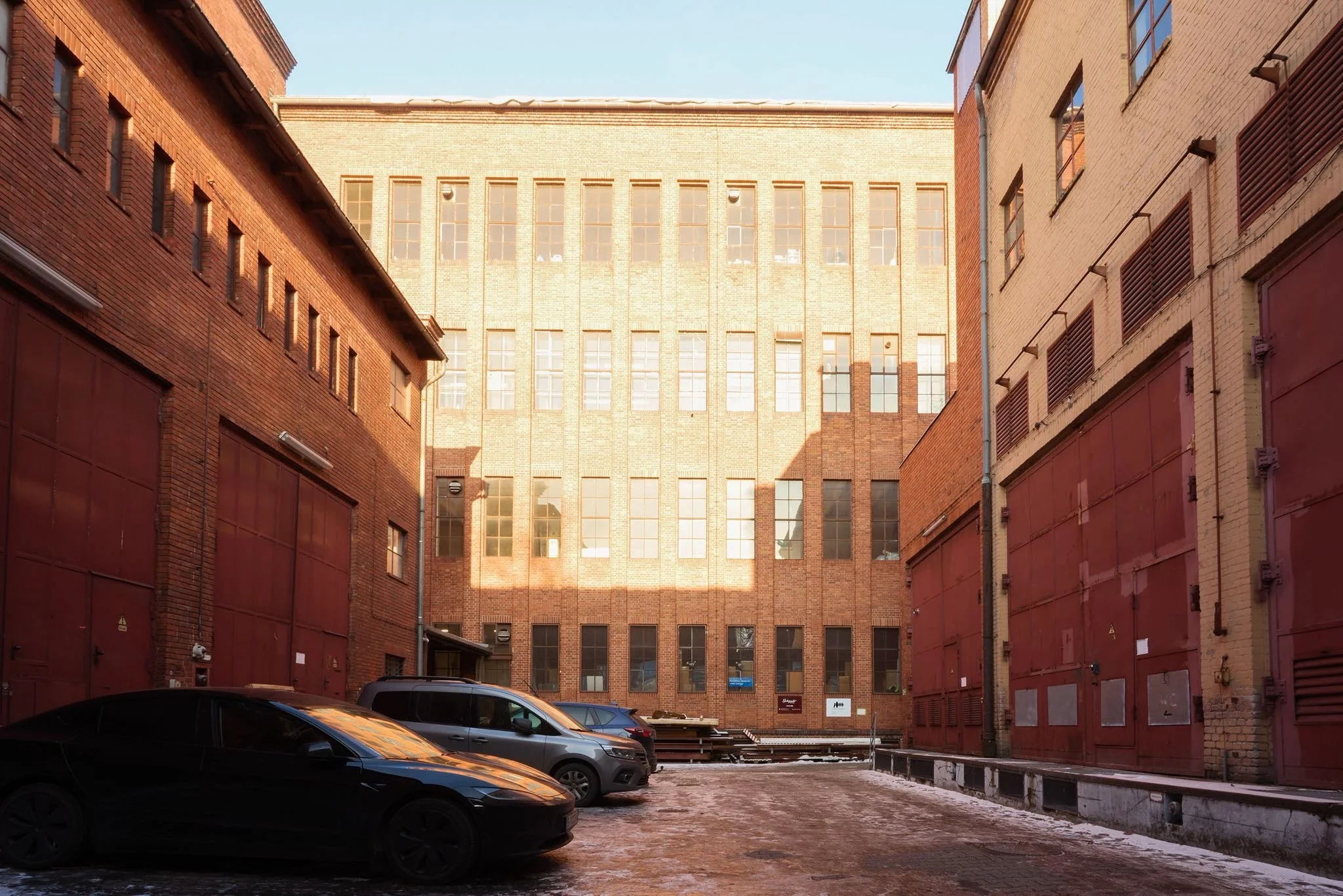 View of an urban parking lot with several cars parked in front of tall brick and concrete buildings, some of which have red roller doors and windows.