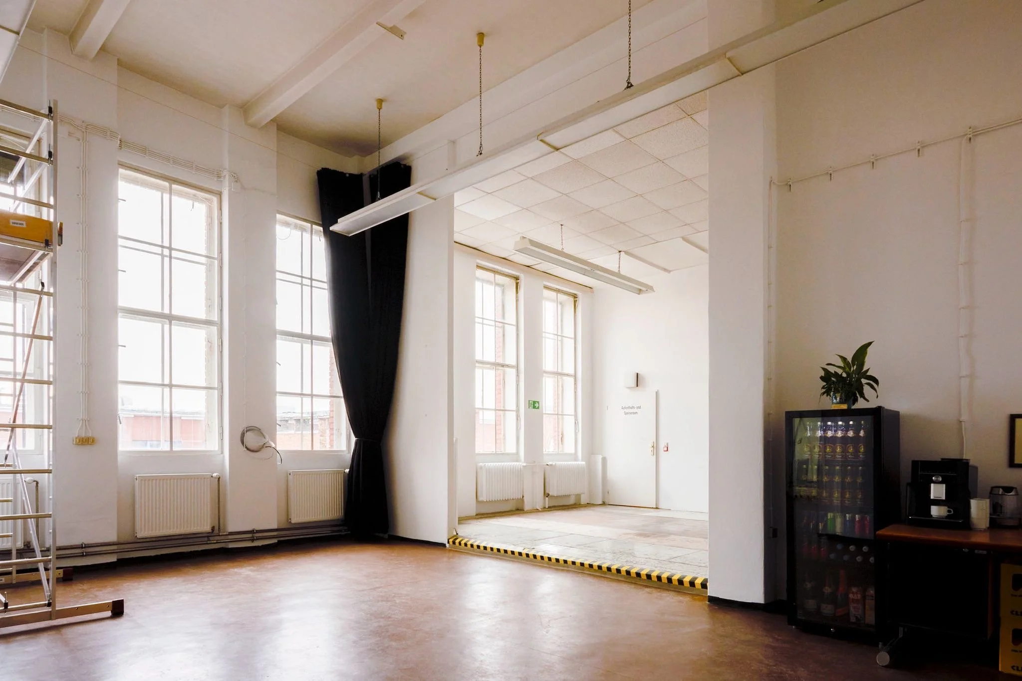 An empty room with large windows letting in natural light, white walls, a black curtain, a wine cooler, a potted plant, and a ladder on the left side.