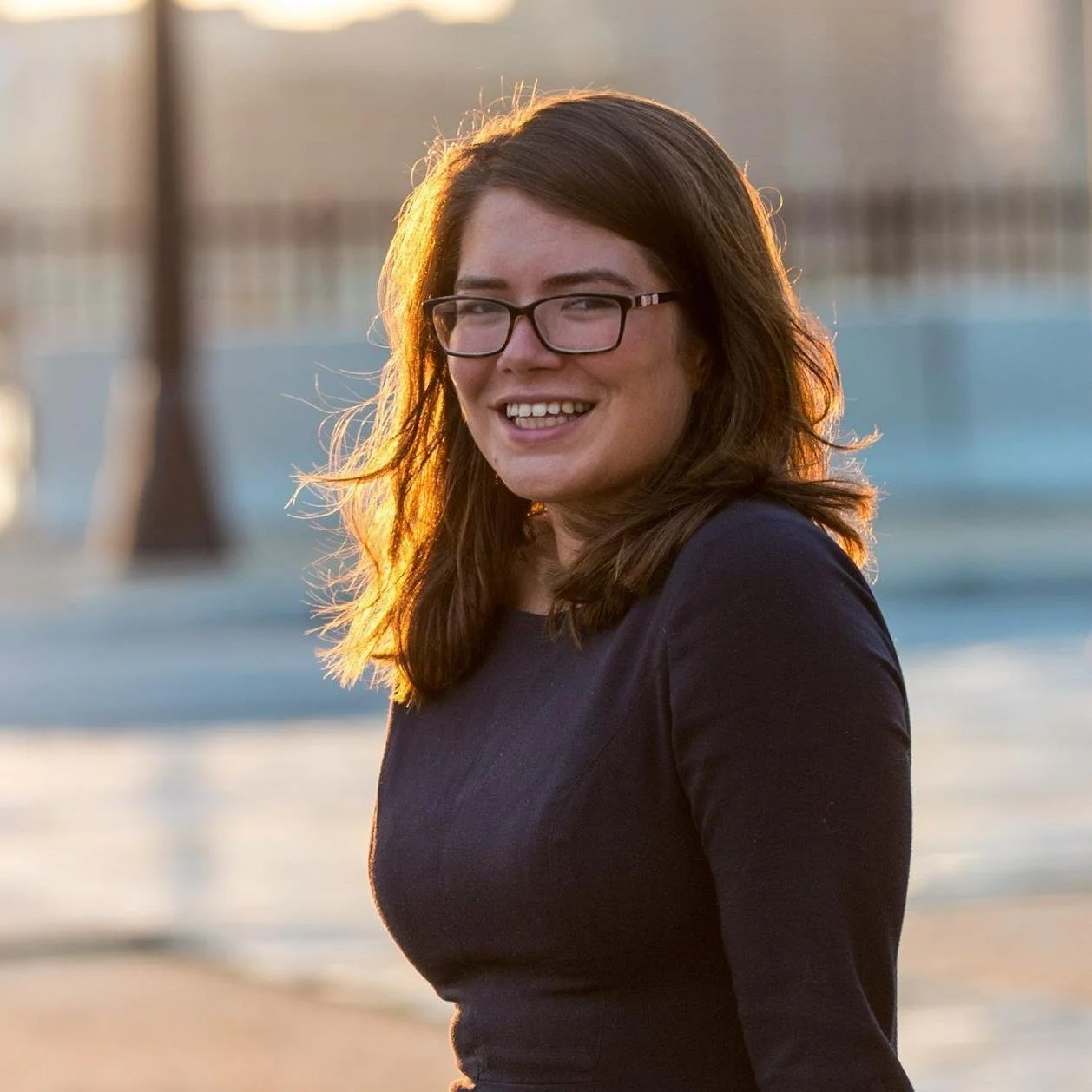 A white woman with straight brown hair and glasses smiles. The sun is setting and glows off her hair.