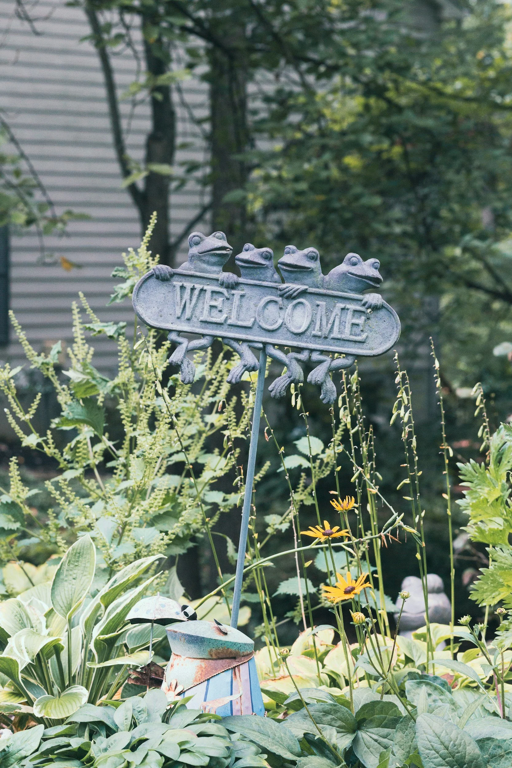 A garden sign featuring four frog figures with the word "WELCOME" surrounded by various plants and flowers.