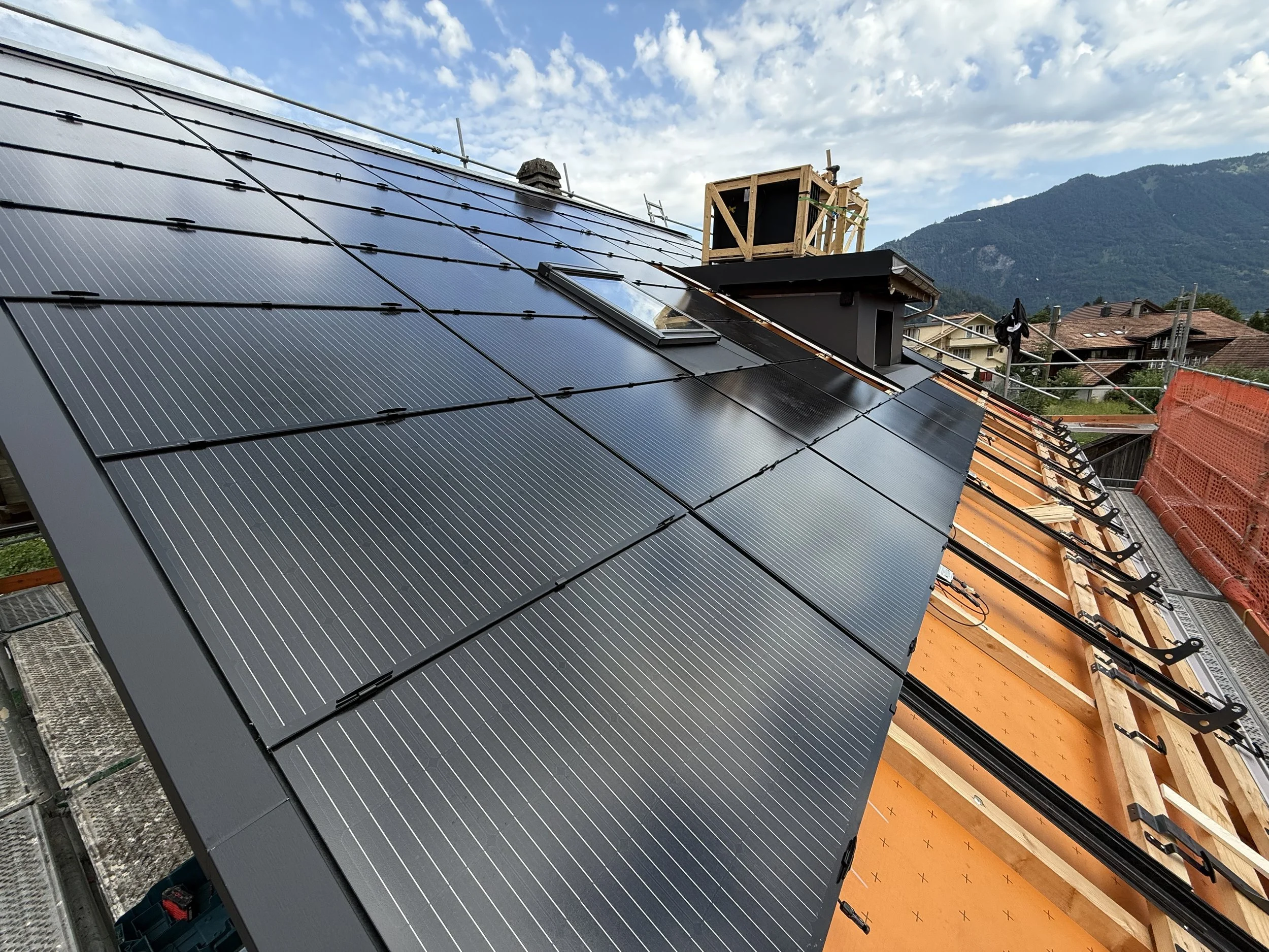 Dach mit schwarzen Solarmodulen auf einer Baustelle, im Hintergrund Berge und ein Himmel mit Wolken.
