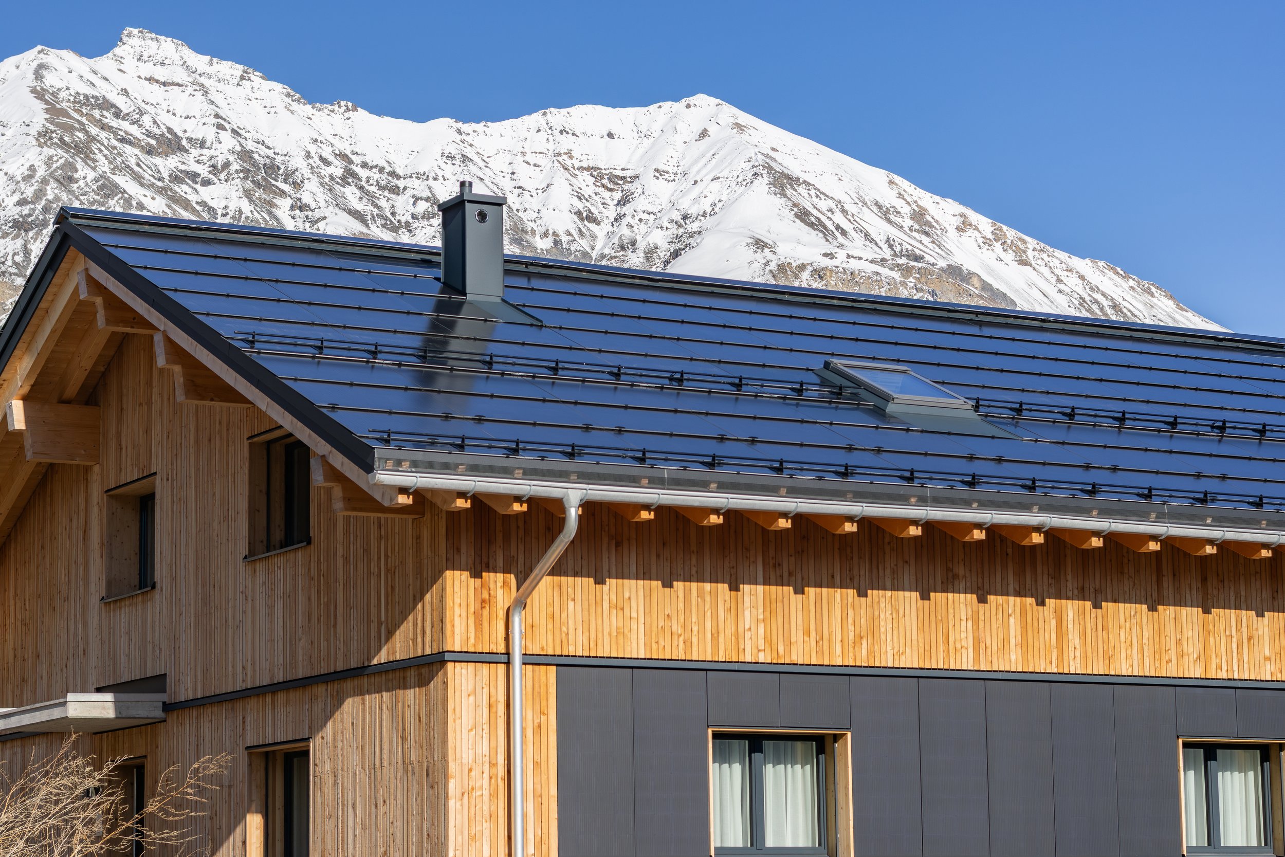 Holzhaus mit blauer Metallschindeldach vor schneebedeckten Bergen unter blauem Himmel.