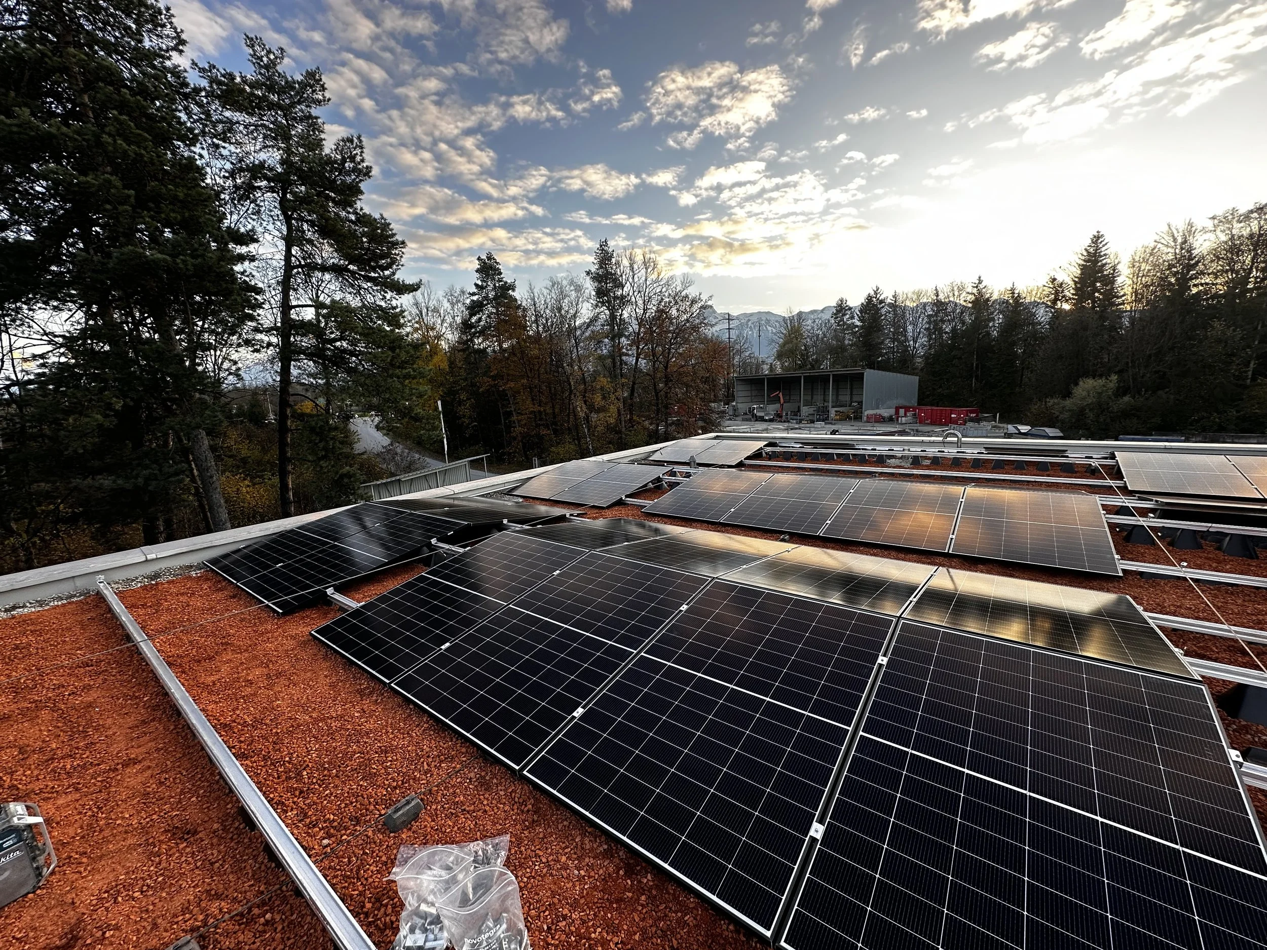 Photovoltaikanlage auf einem roten Flachdach vor einem Himmel mit Wolken, umgeben von Bäumen und Bergen im Hintergrund.