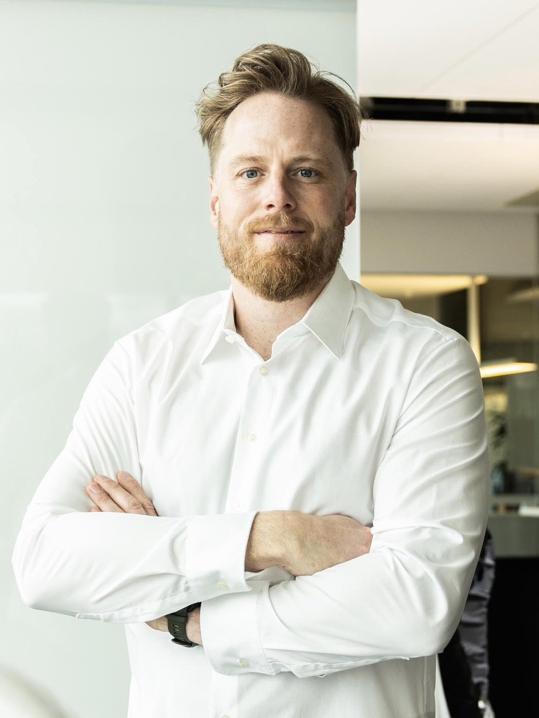 Porträt eines Mannes mit blondem Haar und Bart, der in einem weißen Hemd mit verschränkten Armen in einem modernen Büro steht.