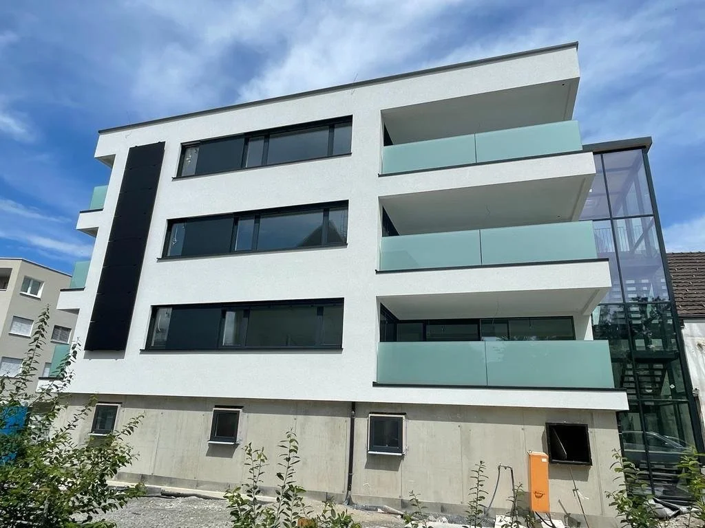 Modernes, weißes Mehrfamilienhaus mit Balkonverglasung und einer seitlichen Glaskonstruktion, im Bau befindlich.