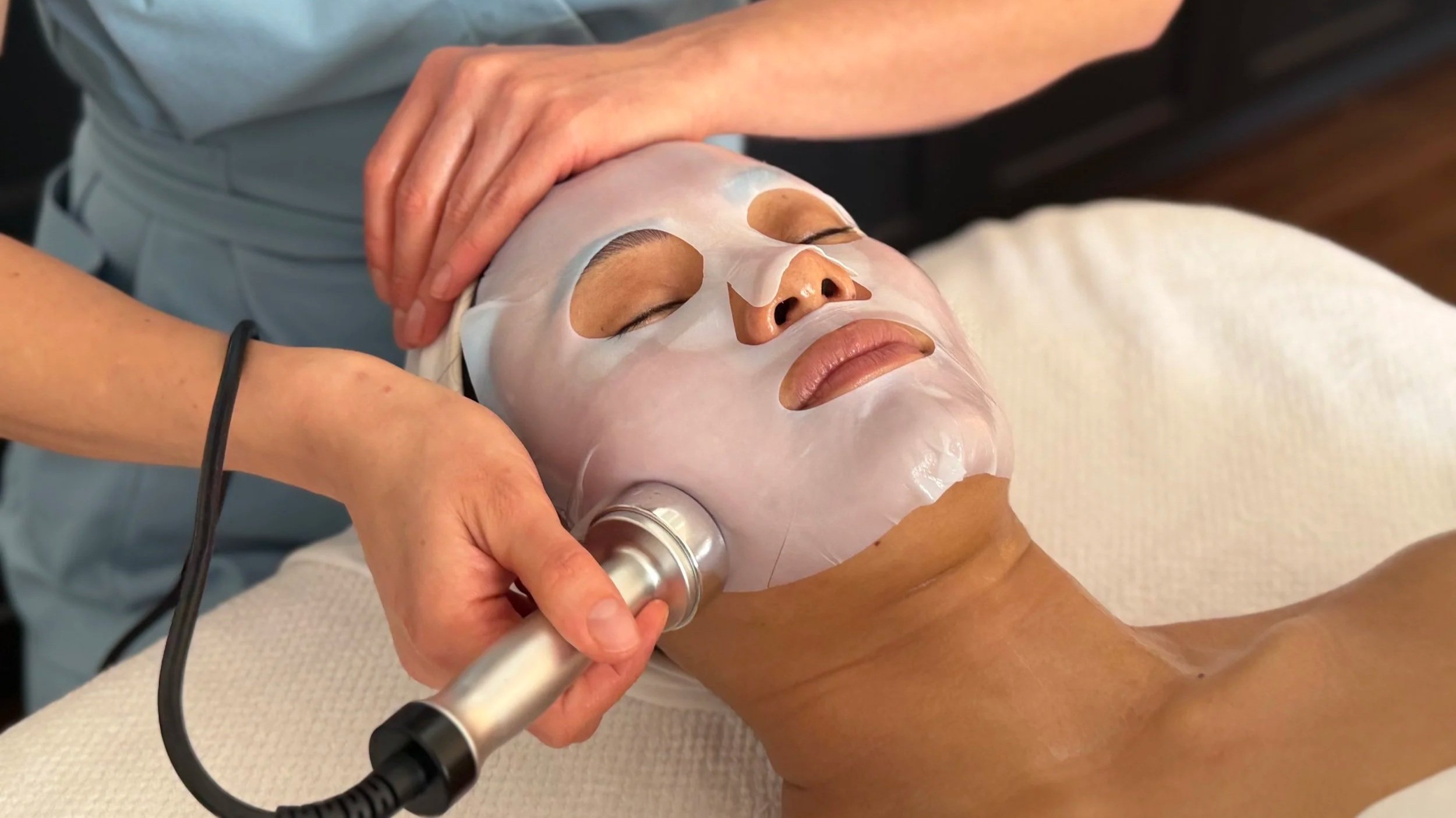 A person receiving a facial treatment with a white sheet mask, while a skincare professional uses a wireless device on their face.