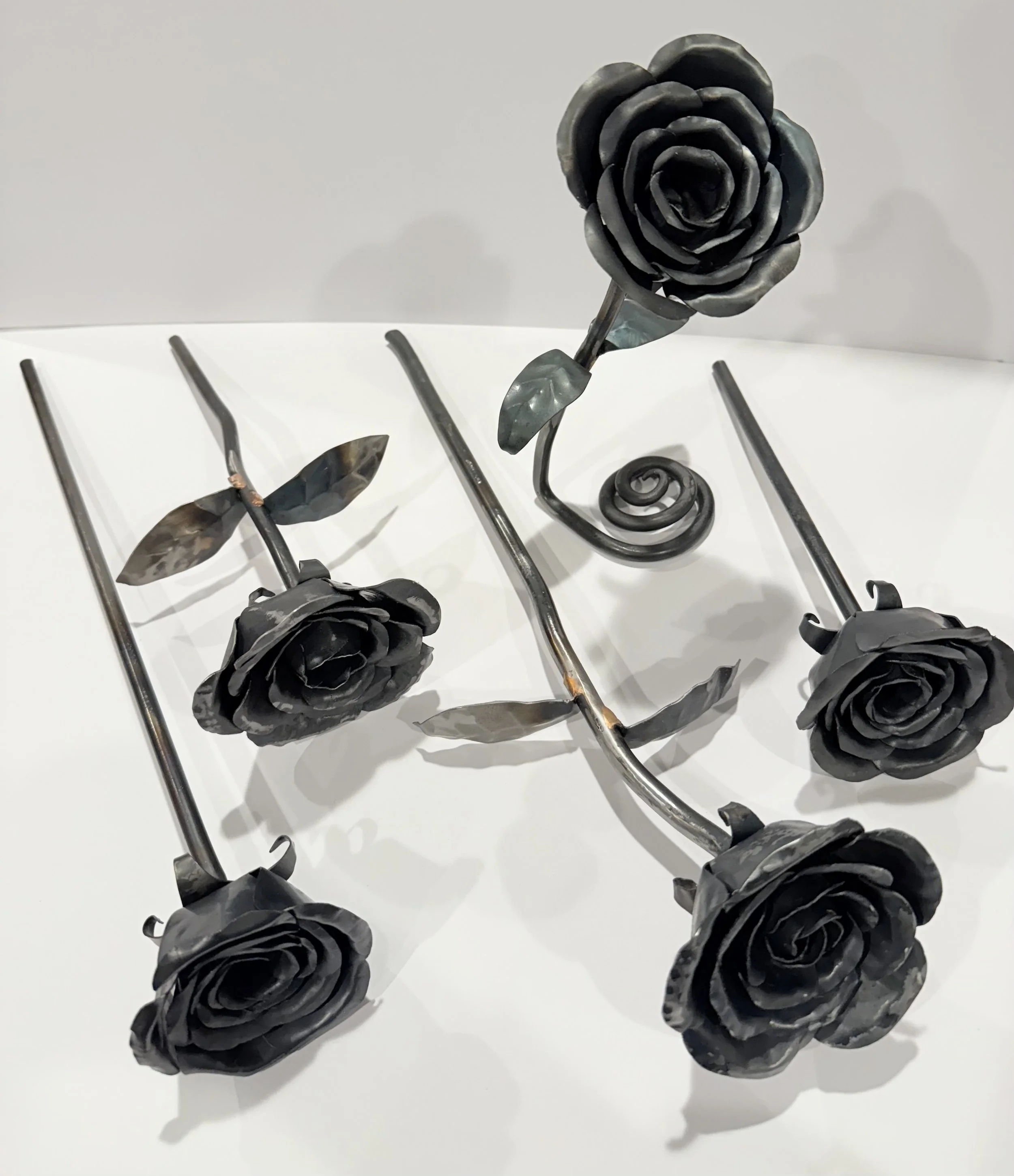 Metal sculpture of five black roses with long stems and leaves, arranged on a white surface.