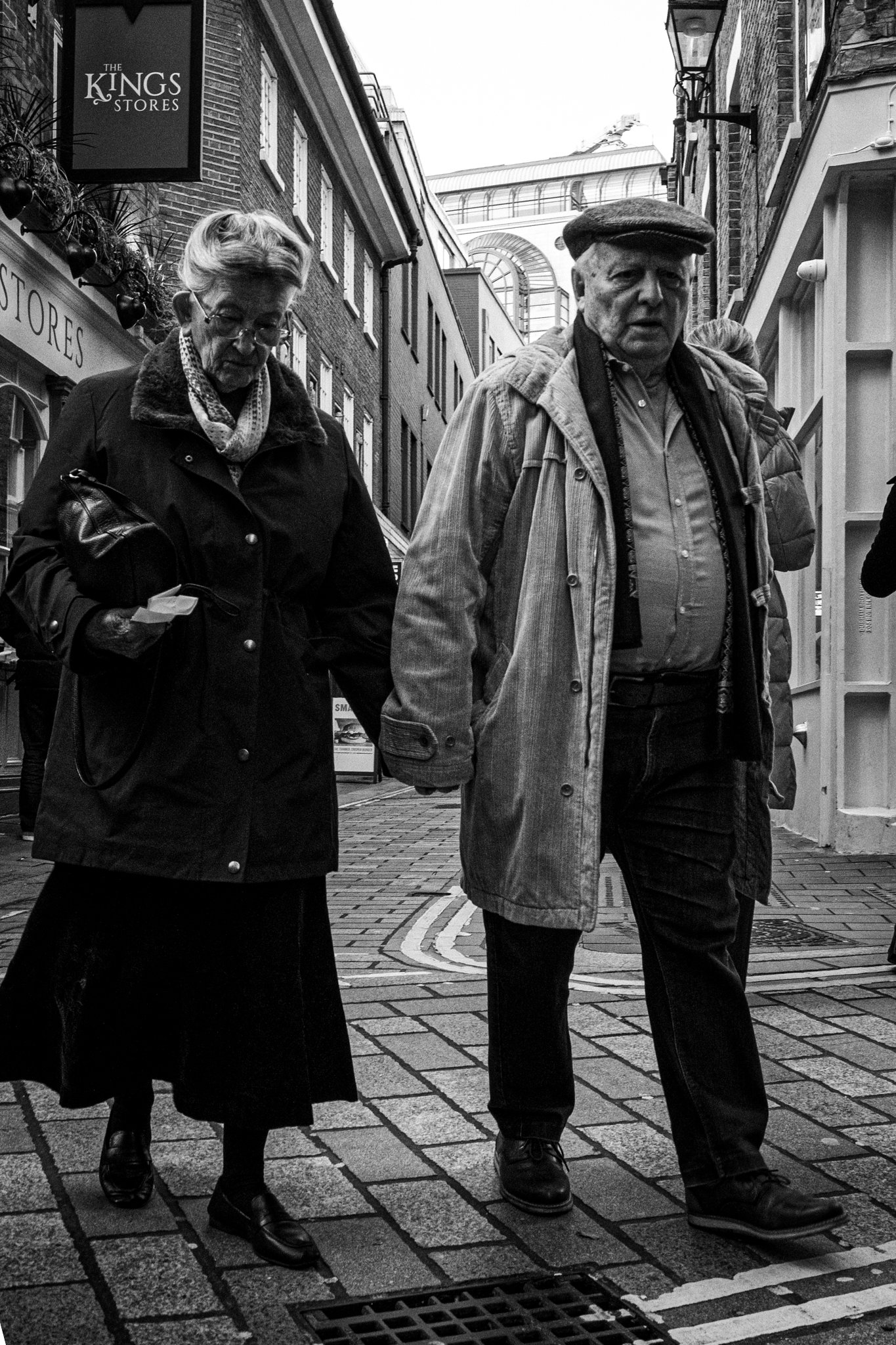 Black and white photo of an elderly couple walking down a narrow city street holding hands. The woman has short hair, glasses, and is wearing a dark coat and skirt. The man has a cap, scarf, light jacket, and dark pants. Buildings and signs are visib