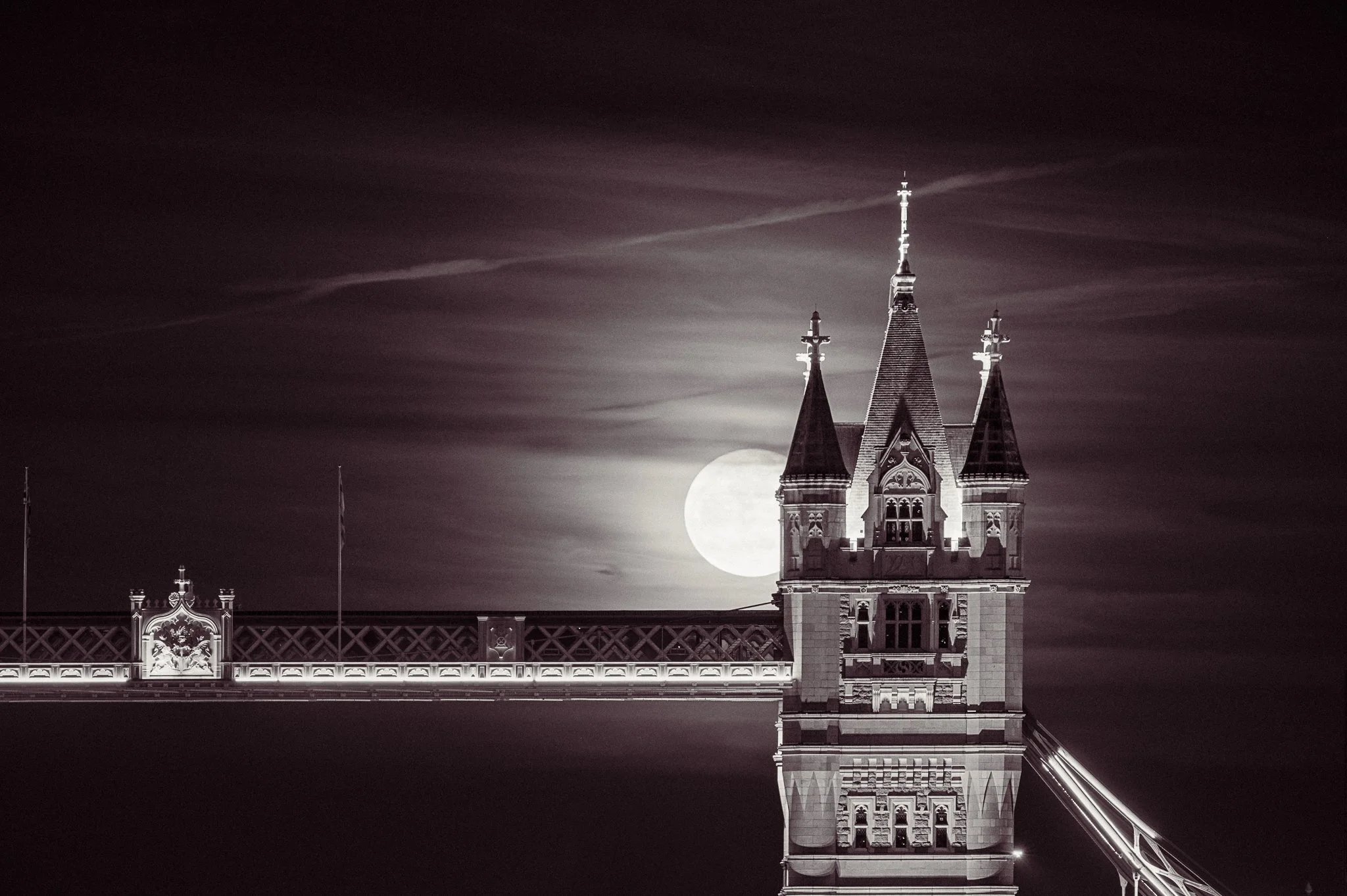 A black-and-white photo of a castle-like building with a tall tower at night, with the moon behind the tower and clouds in the sky.