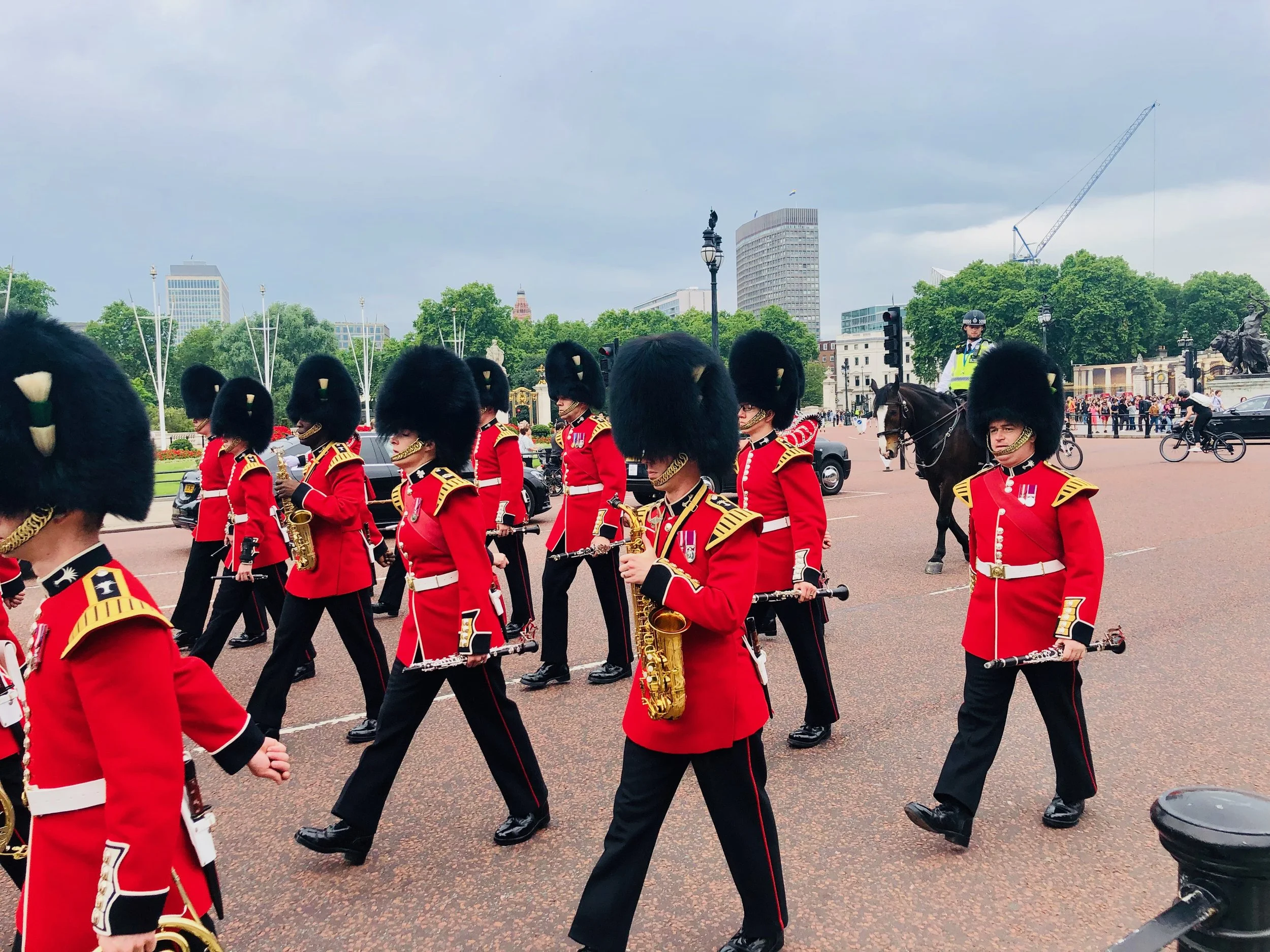 British royal guards marching in ceremonial uniform with tall bearskin hats in a park area with city buildings and trees in the background.