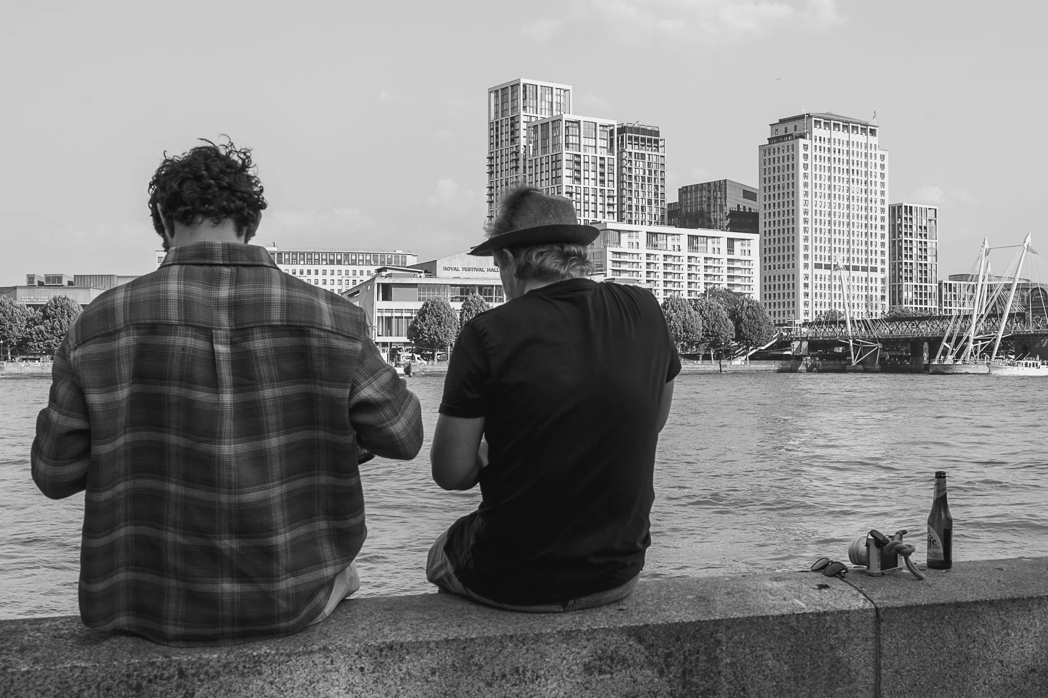 Two men sitting by a river with a city skyline in the background, some items like sunglasses, a water bottle, and a rope are visible on the ground near them.