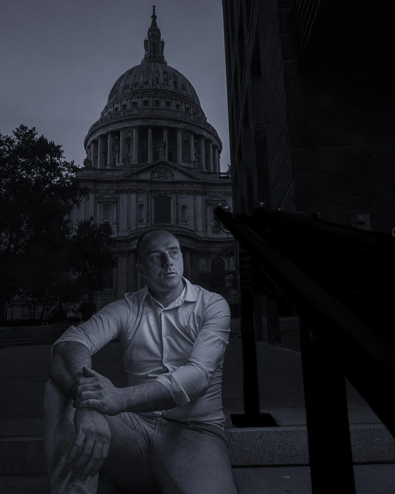 A black and white photo showing St. Paul's Cathedral, London in the background, taken at night. In the foreground, a person with short hair, wearing a collared shirt, sits with their arms crossed, looking slightly up and to the right.