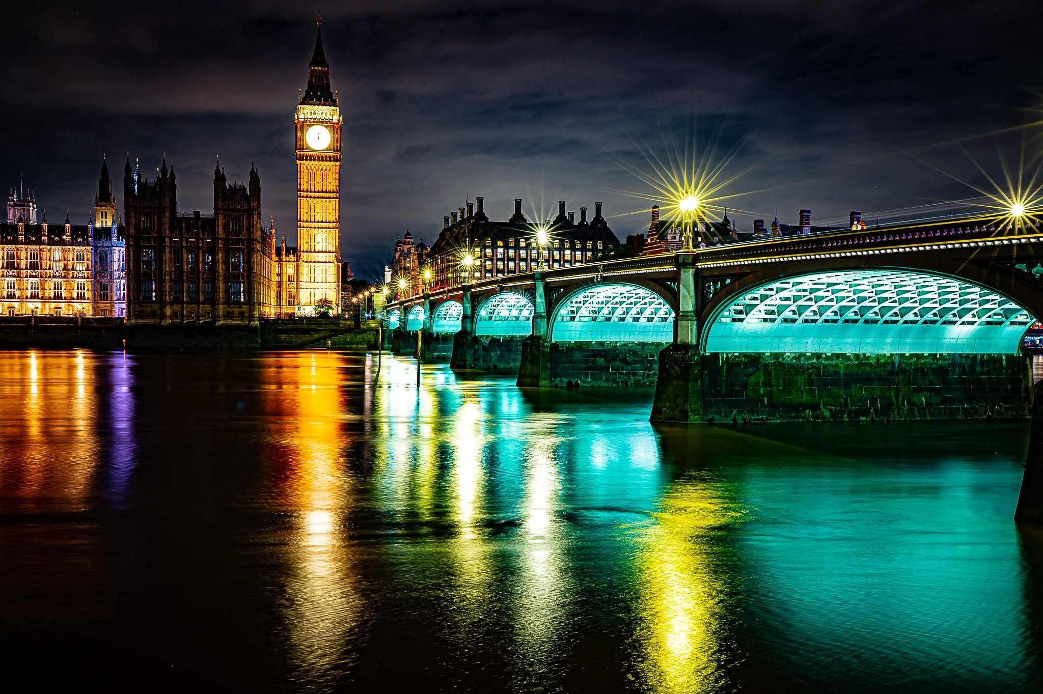 Nighttime view of Big Ben clock tower and Houses of Parliament in London, illuminated with colorful lights, reflected on the river, with a cloudy sky overhead.