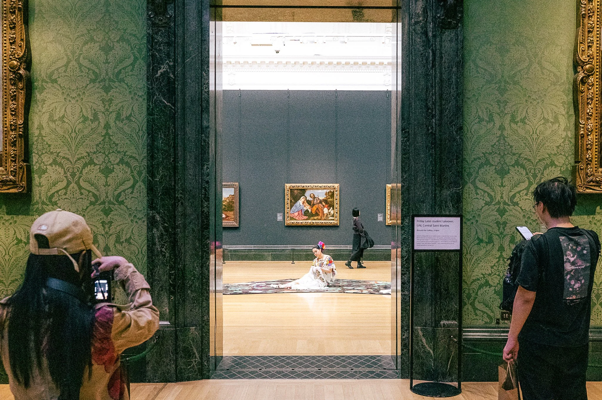 People viewing art in a museum with a performer dressed in traditional attire sitting on the floor in front of paintings.