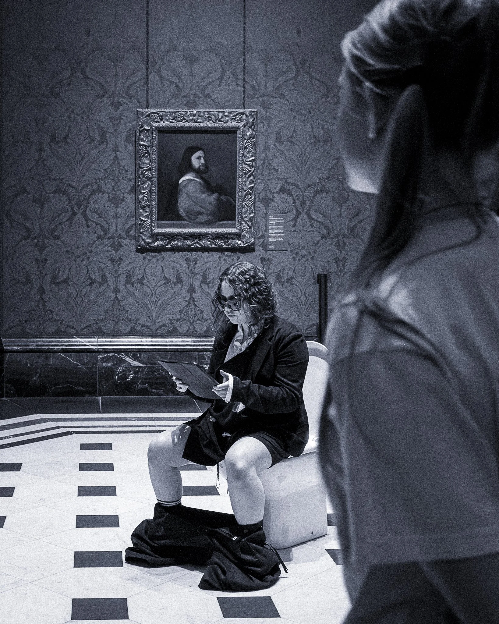 A person with curly hair and glasses sitting on a toilet in a public restroom, reading a book. Another person wearing a dark jacket is partially visible on the right side of the image. There is a framed portrait of Jesus Christ on the wall behind them, with decorative wallpaper.