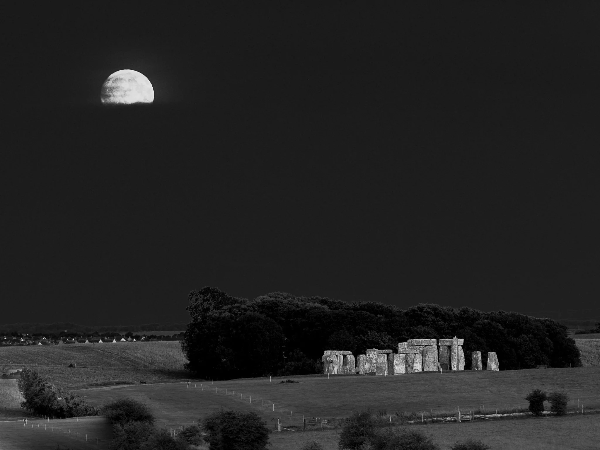 A black and white photo of a landscape at night showing a large moon rising behind a hill, with trees, fields, and a stone fence in the foreground.