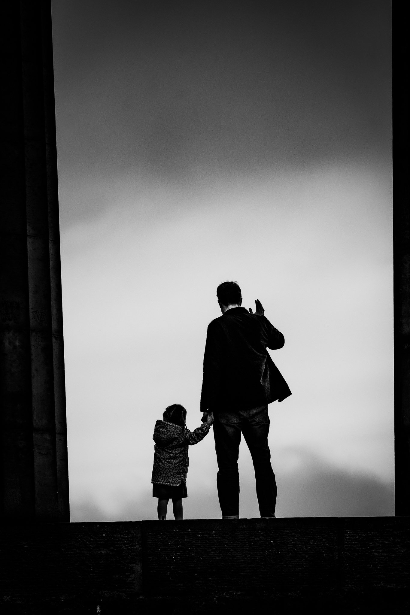 Silhouette of a man and a young girl holding hands against a cloudy sky, viewed from below.