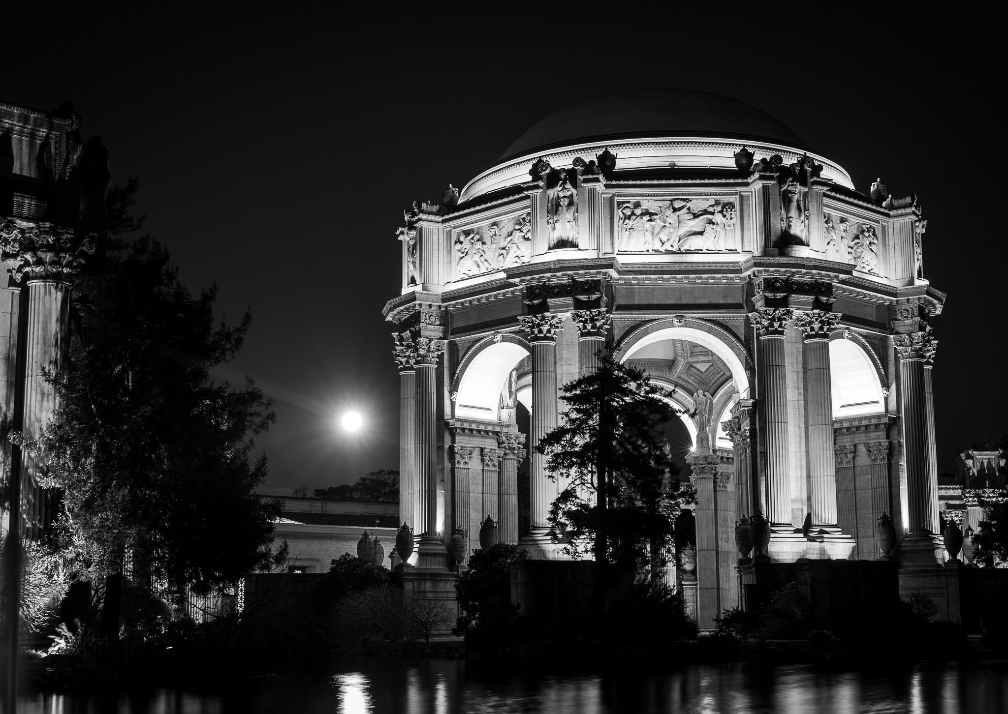 A nighttime view of a classical-style monument with tall columns and elaborate sculpture work, illuminated and reflected in a body of water with silhouetted trees in the foreground.
