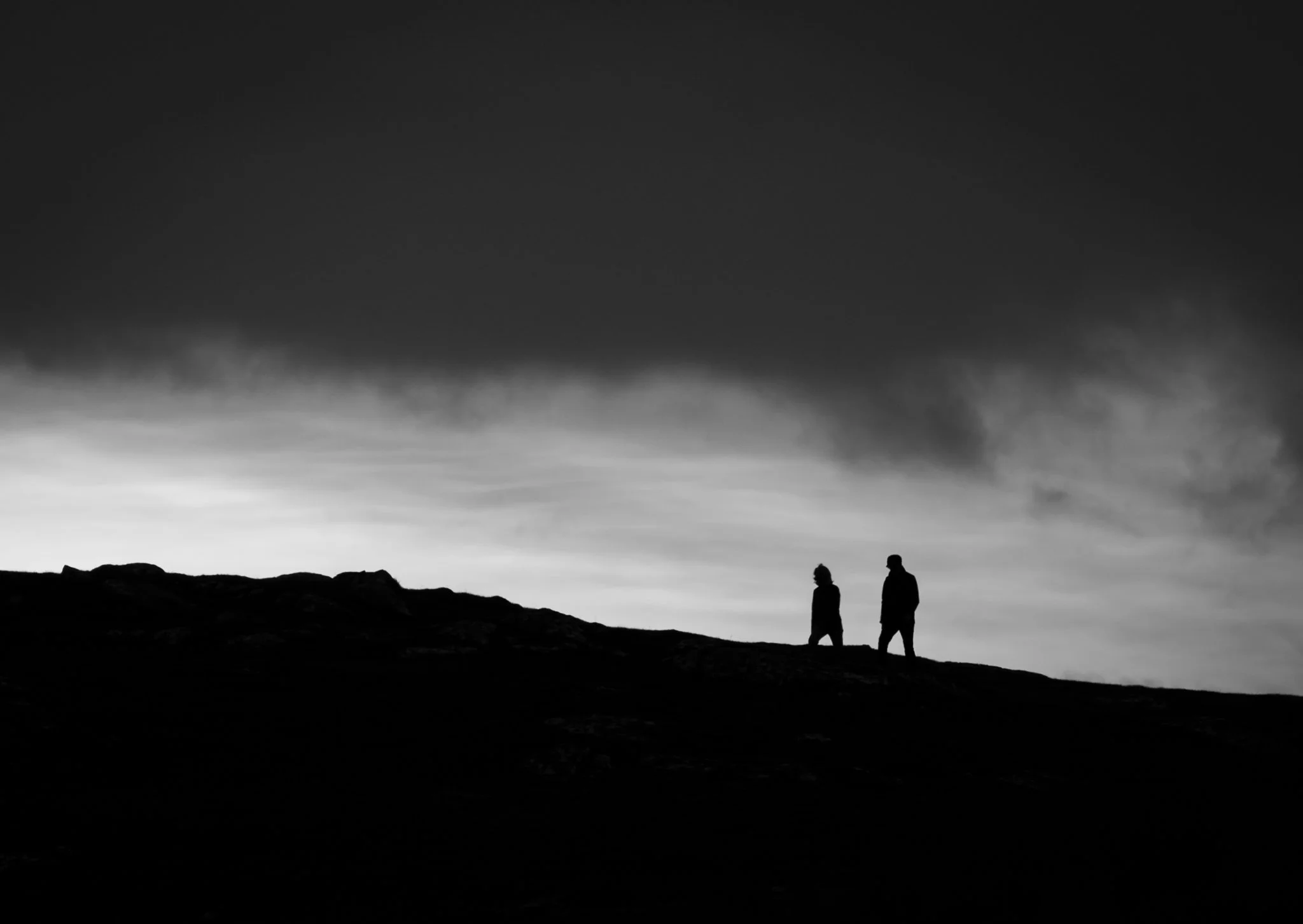 Silhouettes of two people walking on a hill against a cloudy sky at dusk or dawn.