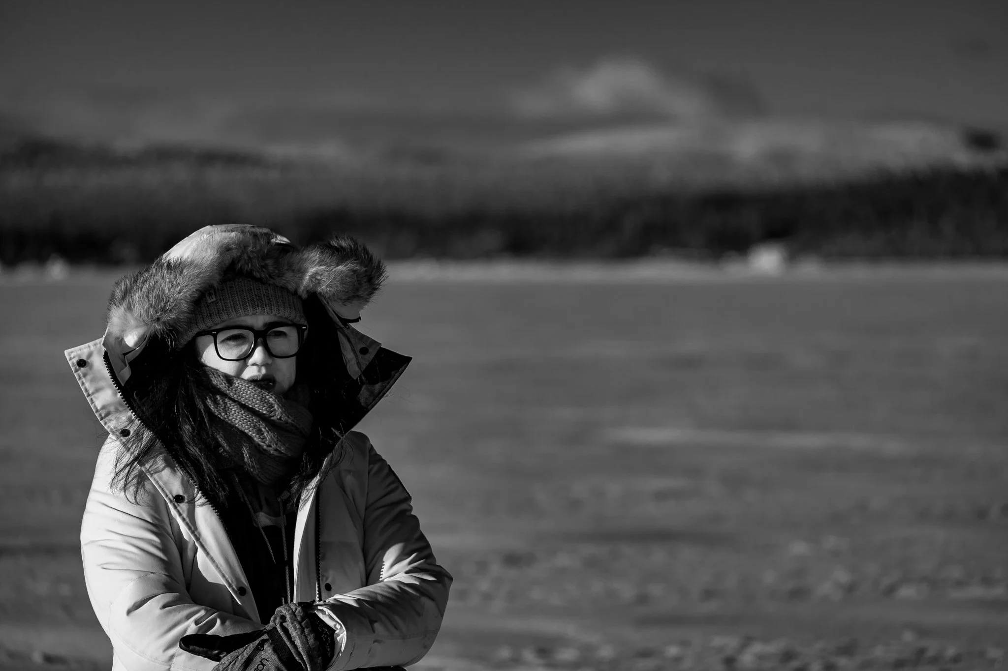 A woman wearing glasses, a winter coat with a fur-lined hood, a knit hat, a scarf, and gloves stands outdoors in a cold environment with a body of water and landscape in the background.