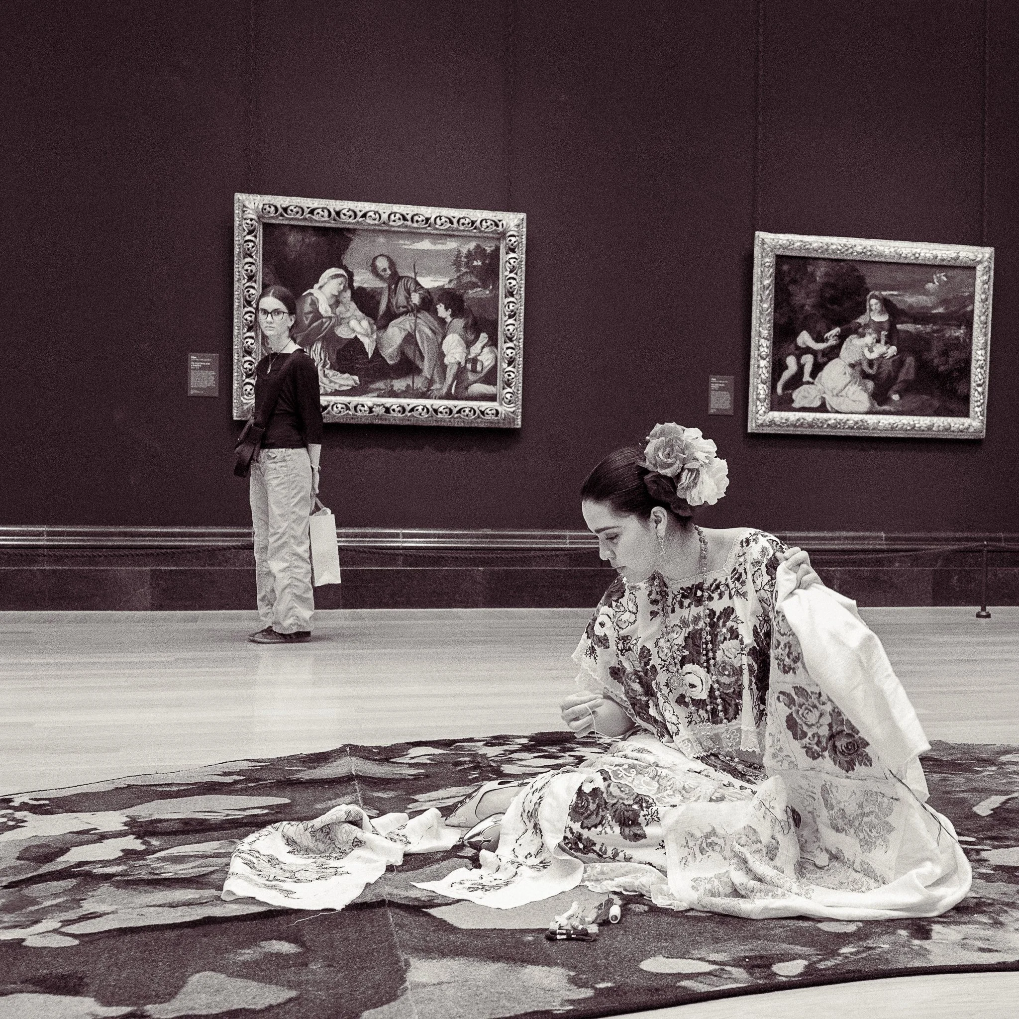 Black and white photo of a woman in a floral dress and flowers in her hair sitting on a large piece of fabric on the floor, with two framed pictures hanging on a dark wall behind her and a woman in glasses standing nearby.