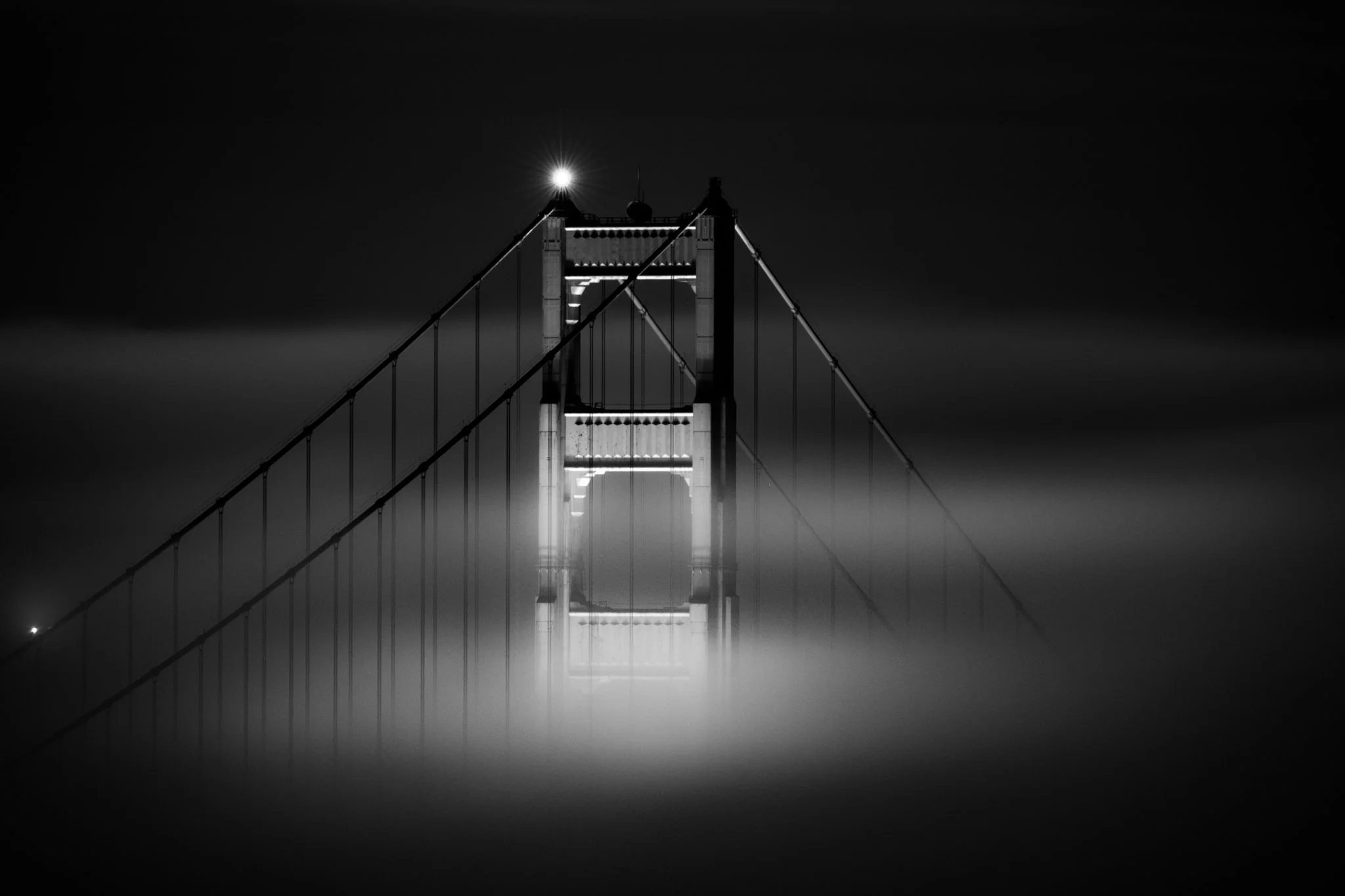 Black-and-white photo of the Golden Gate Bridge at night shrouded in fog with illumination from a light on the tower